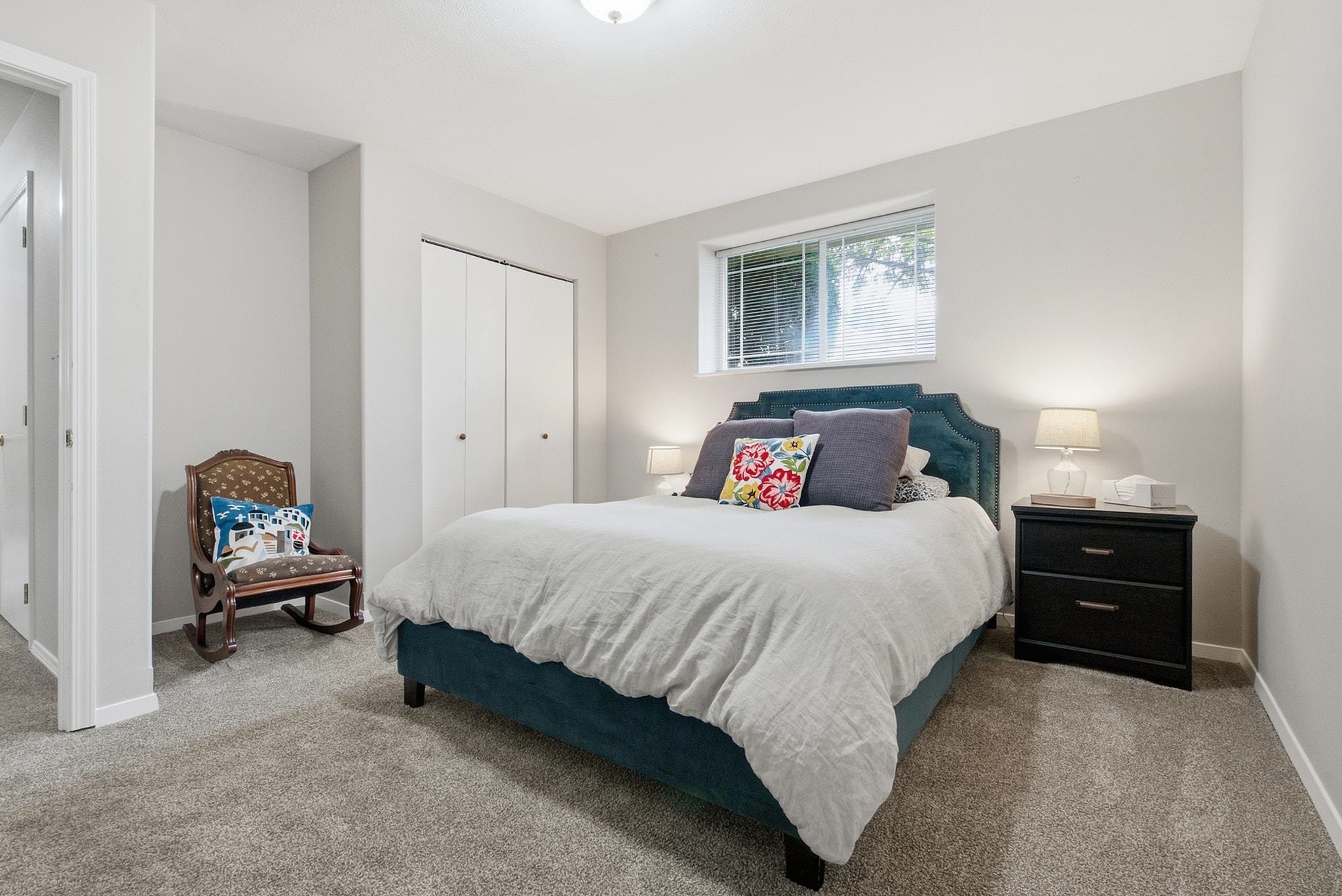 Furnished bedroom with a teal upholstered bed, colorful pillows, carpet, and a window with blinds.