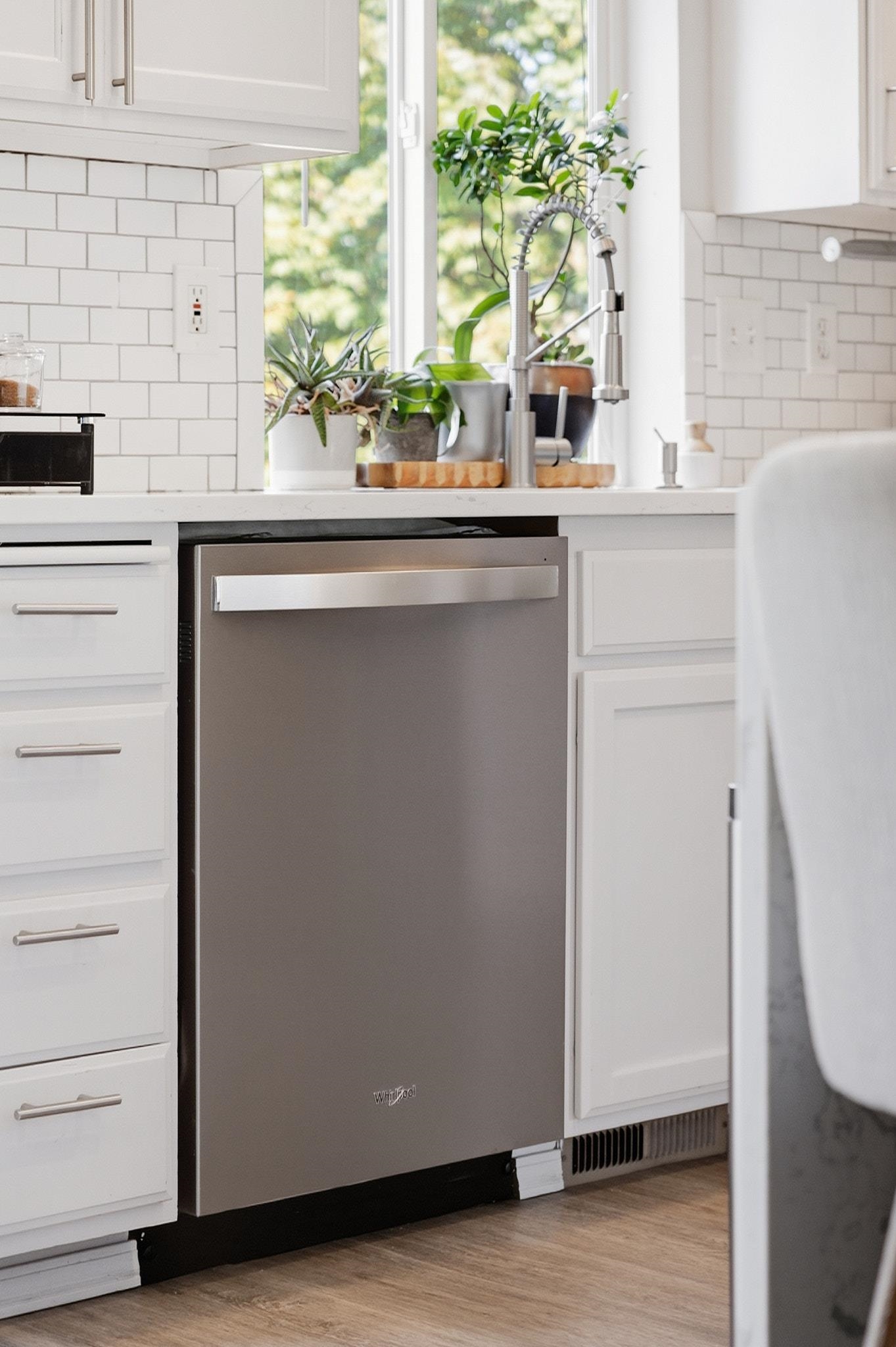 Kitchen with a stainless steel dishwasher, white cabinets, subway tile backsplash, and plants by the window.