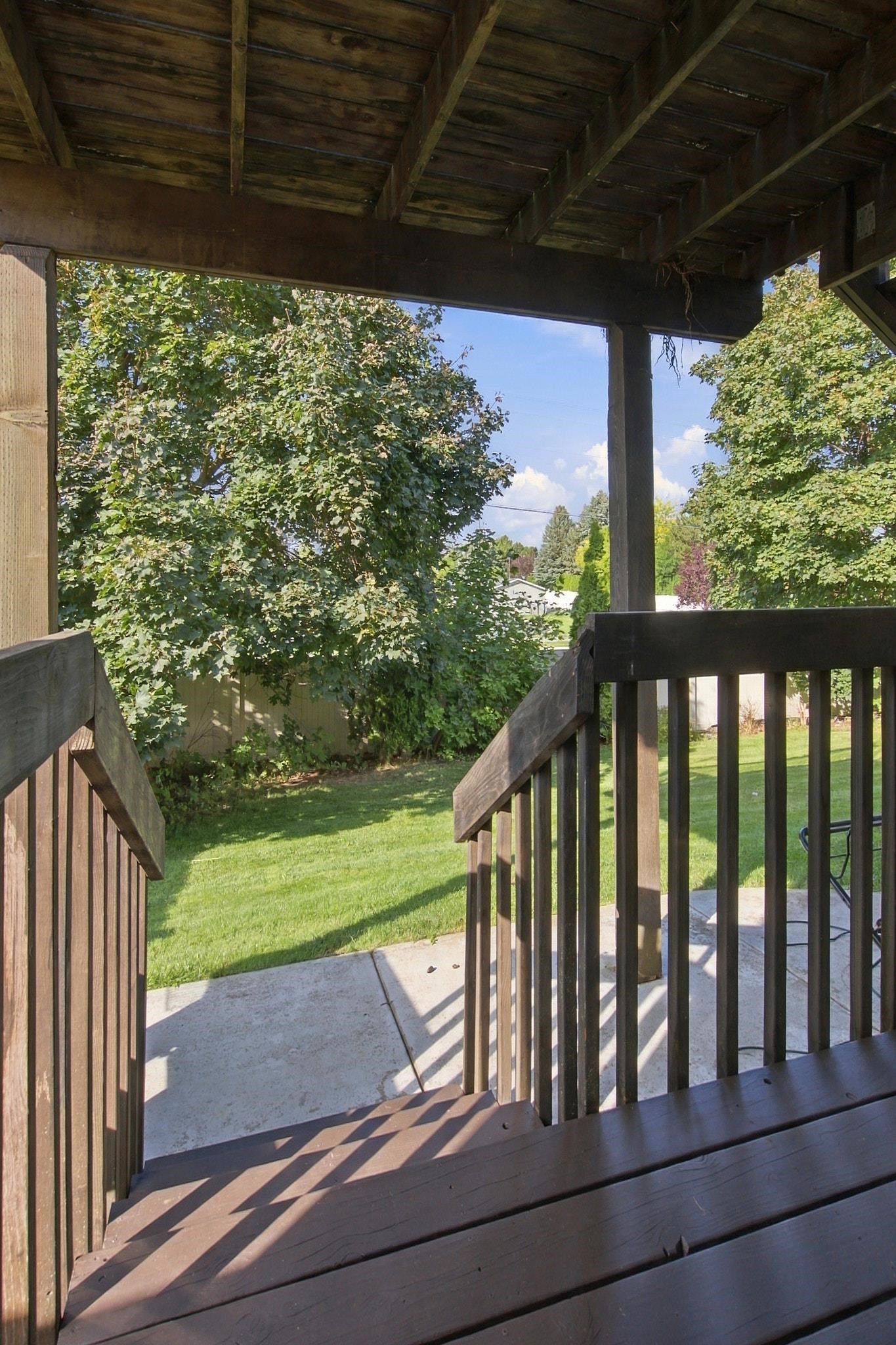 Covered wooden deck overlooking a green lawn and mature trees, with stairs leading to a concrete patio.
