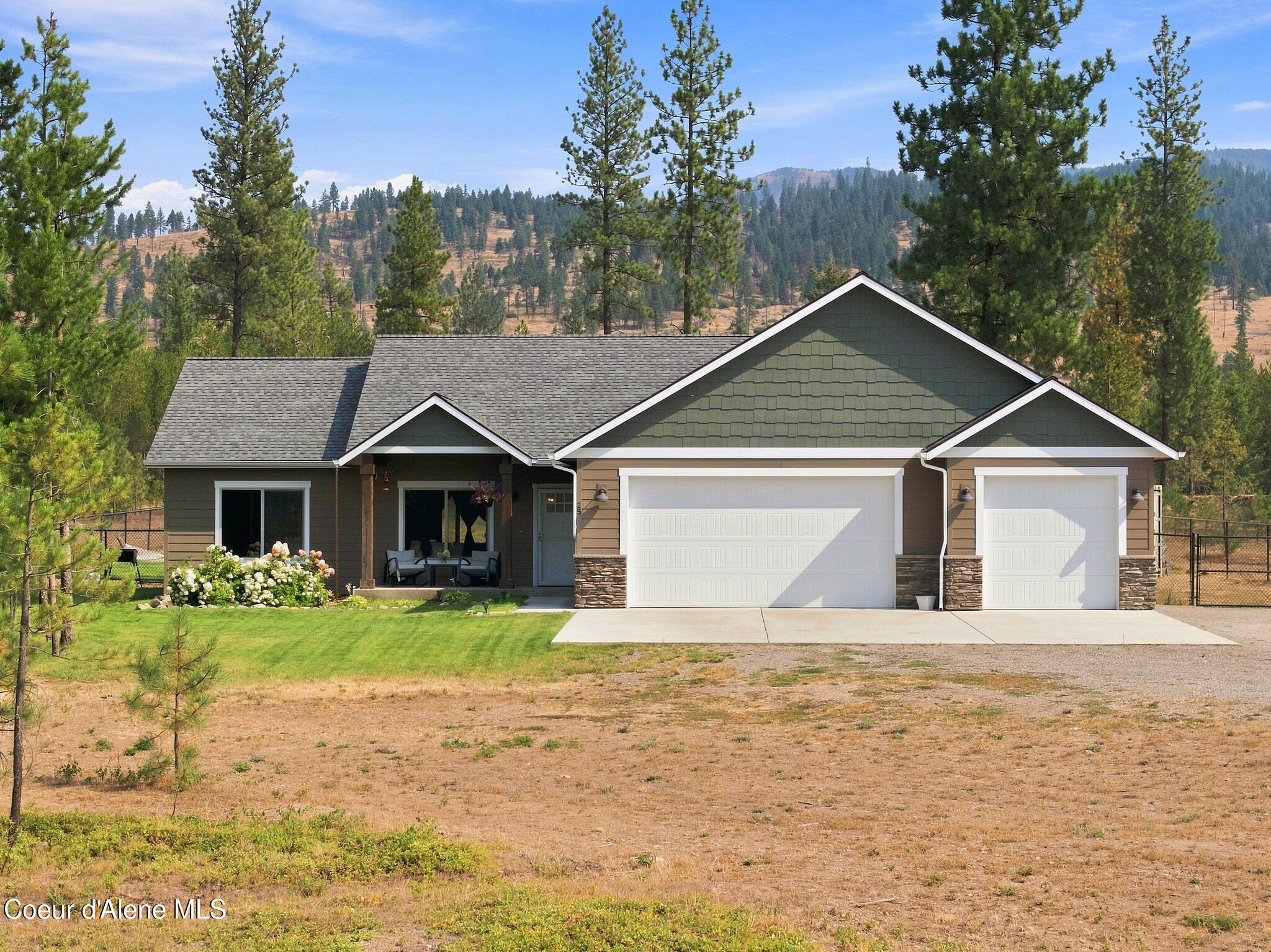 Single-story home with two white garage doors, a covered seating area, and pine-covered hills behind it.