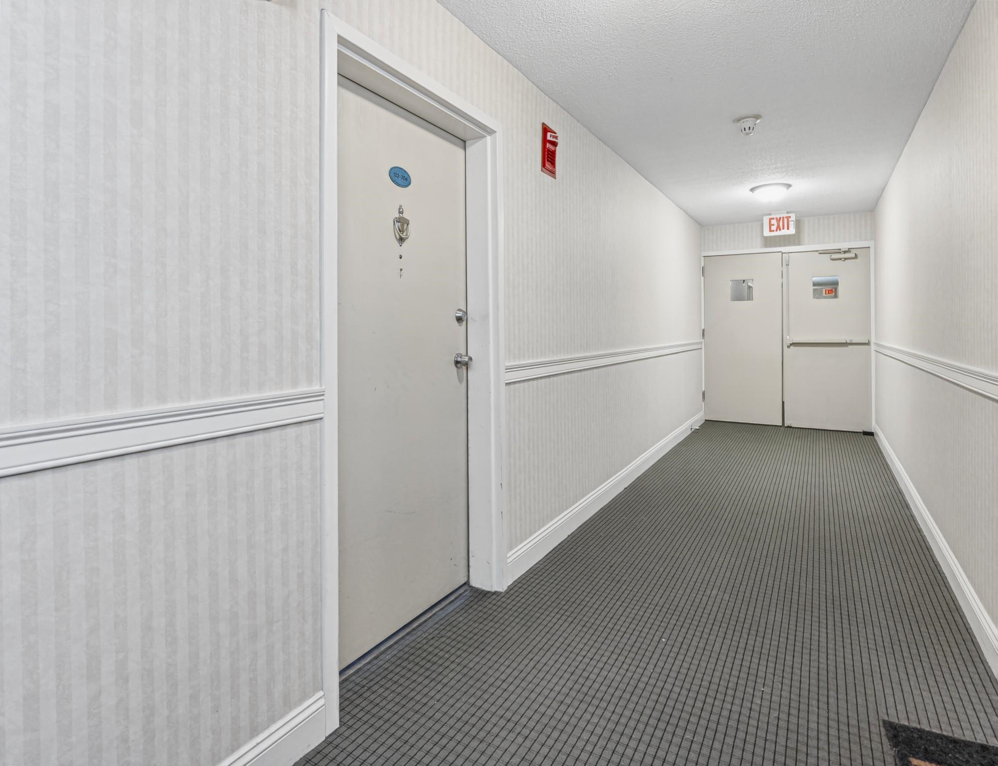 Condominium hallway with a unit door, striped wallpaper, and emergency exit doors at the far end.