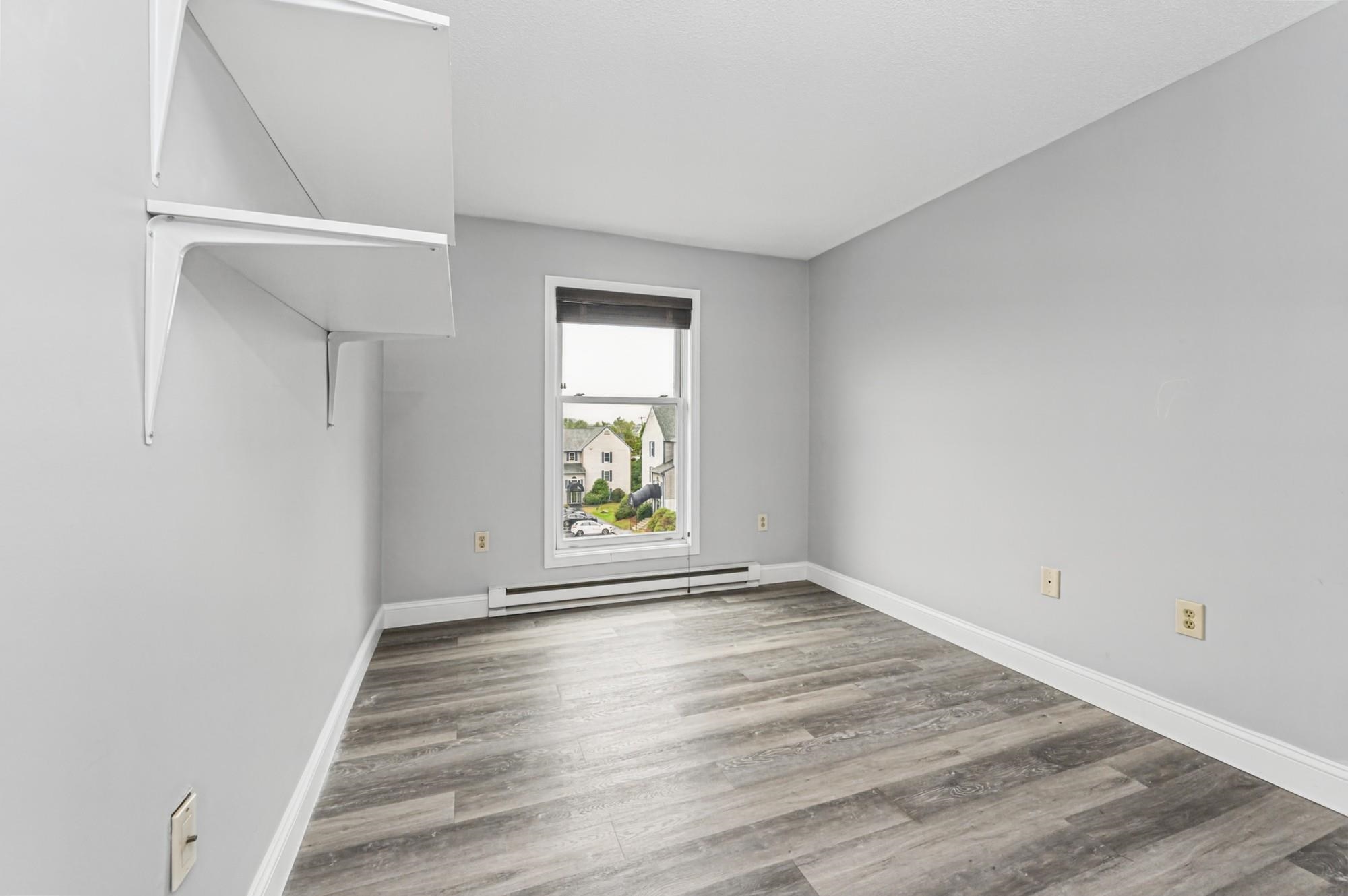 Empty room with gray wood-look flooring, wall-mounted shelves, and a window above a baseboard heater.