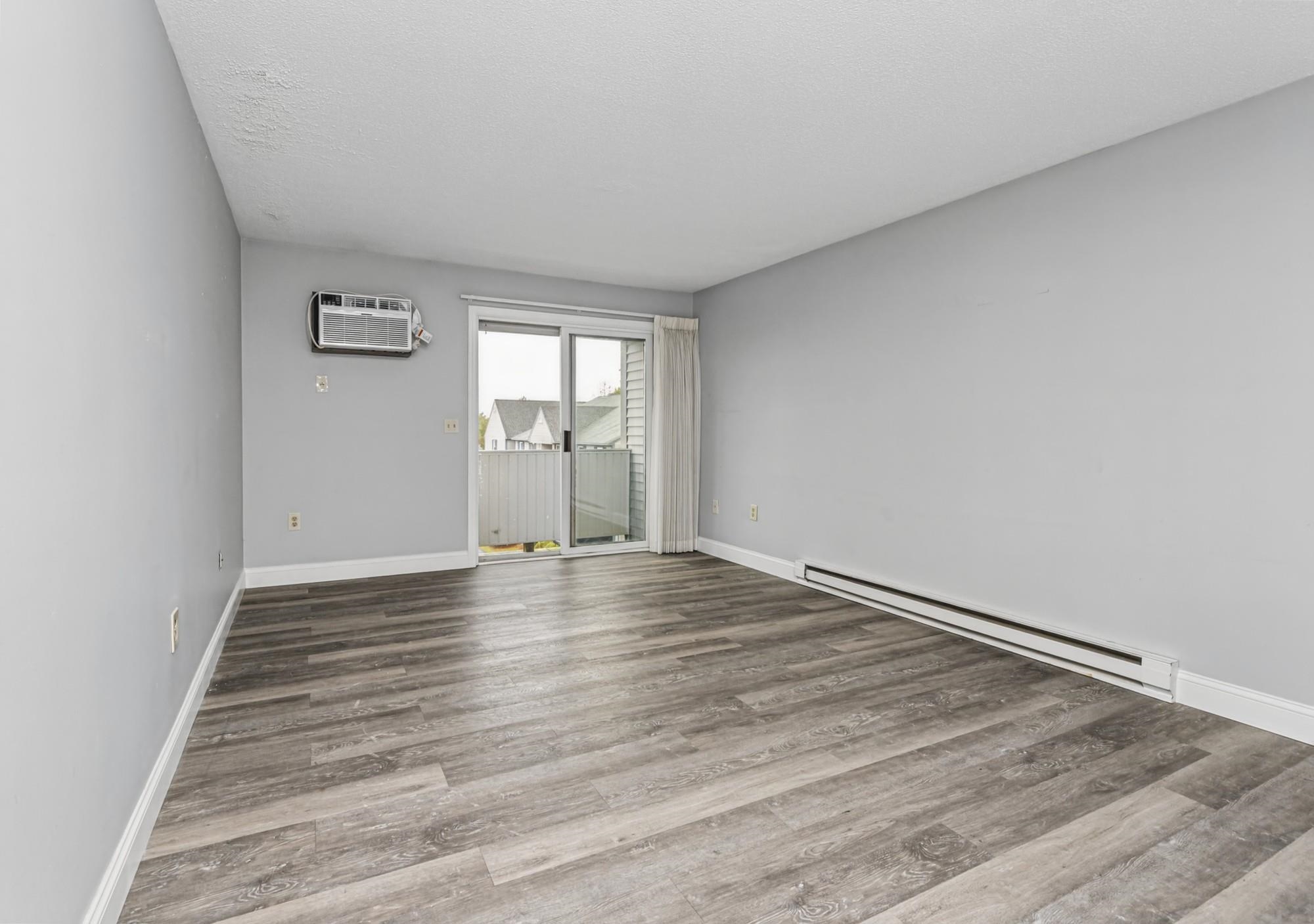 Empty living room with gray wood-look floors, wall air conditioner, and sliding glass balcony door.