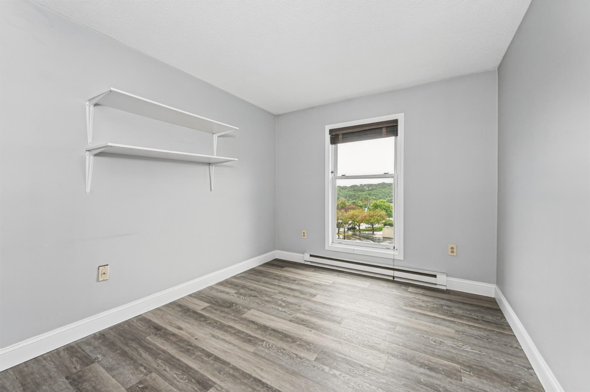 Empty room with gray wood-look flooring, two wall shelves, and a window overlooking trees.
