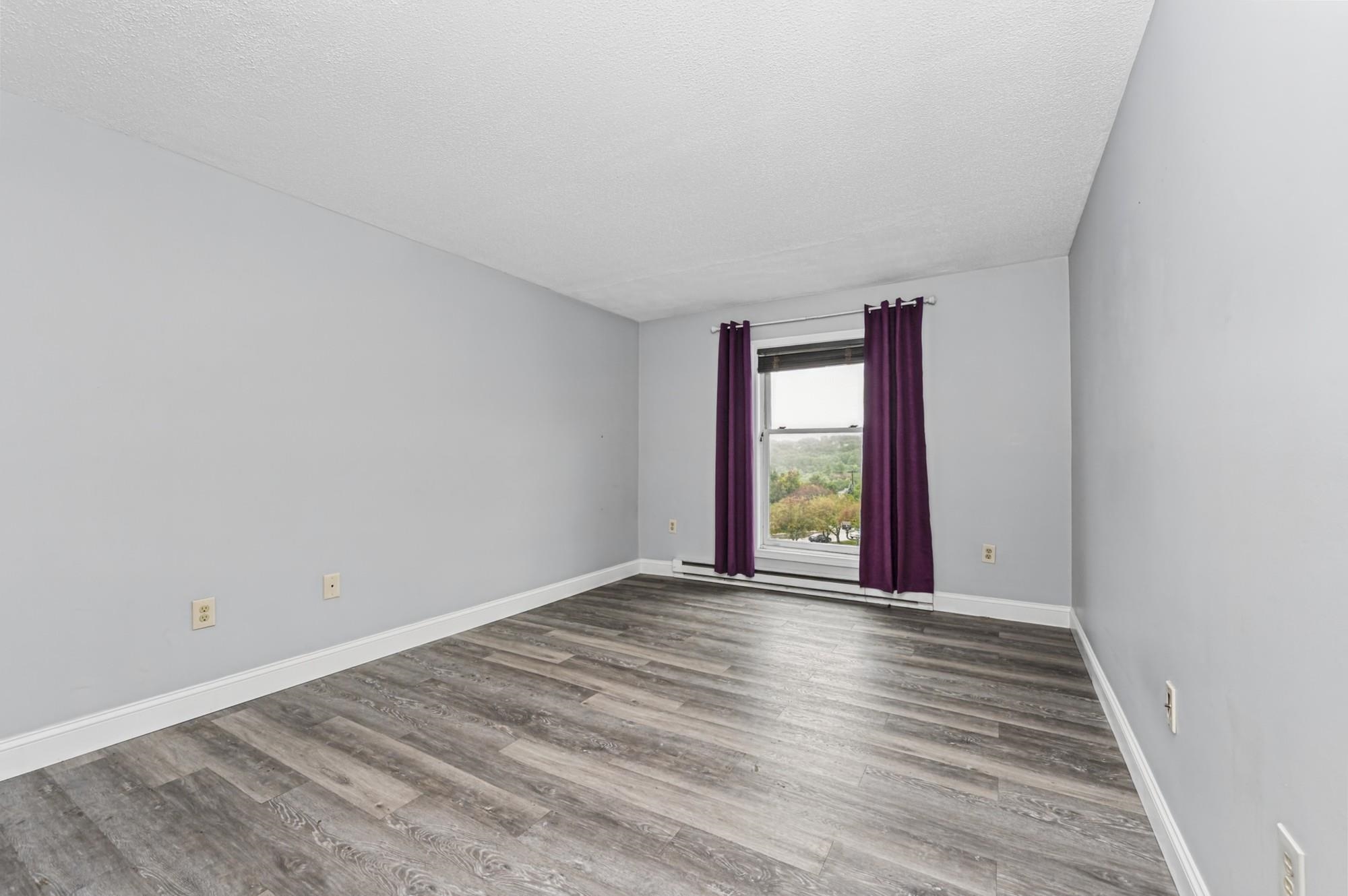 Empty bedroom with gray wood-look flooring and a window framed by purple curtains.