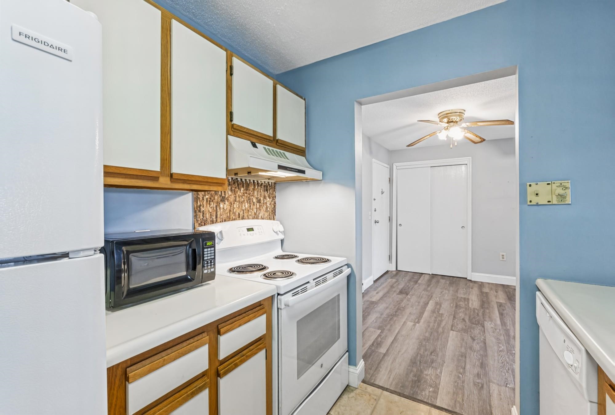 Kitchen with white appliances and wood-trimmed cabinets opening to a gray-floored hallway.