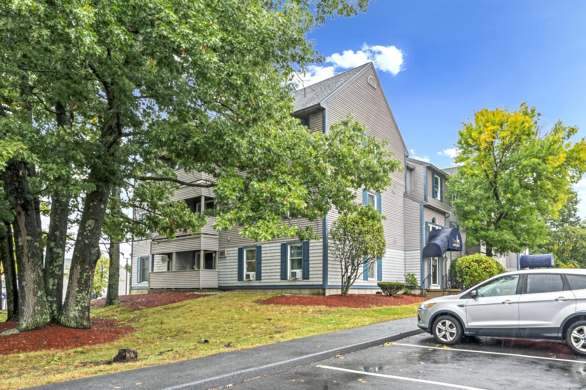 Condominium building with balconies and blue trim, partly hidden by trees beside a parking lot.