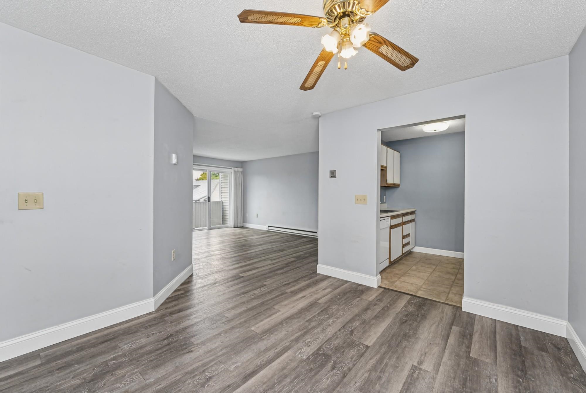 Vacant living area with gray plank flooring, a ceiling fan, sliding glass door, and adjoining kitchen.