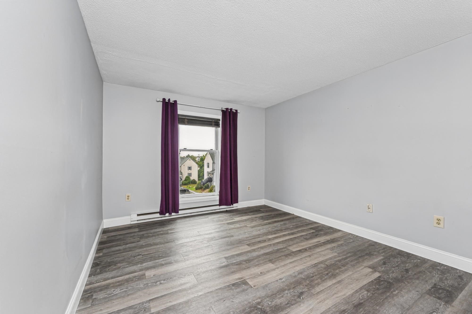 Empty room with gray wood-look flooring and purple curtains around a window overlooking nearby houses.