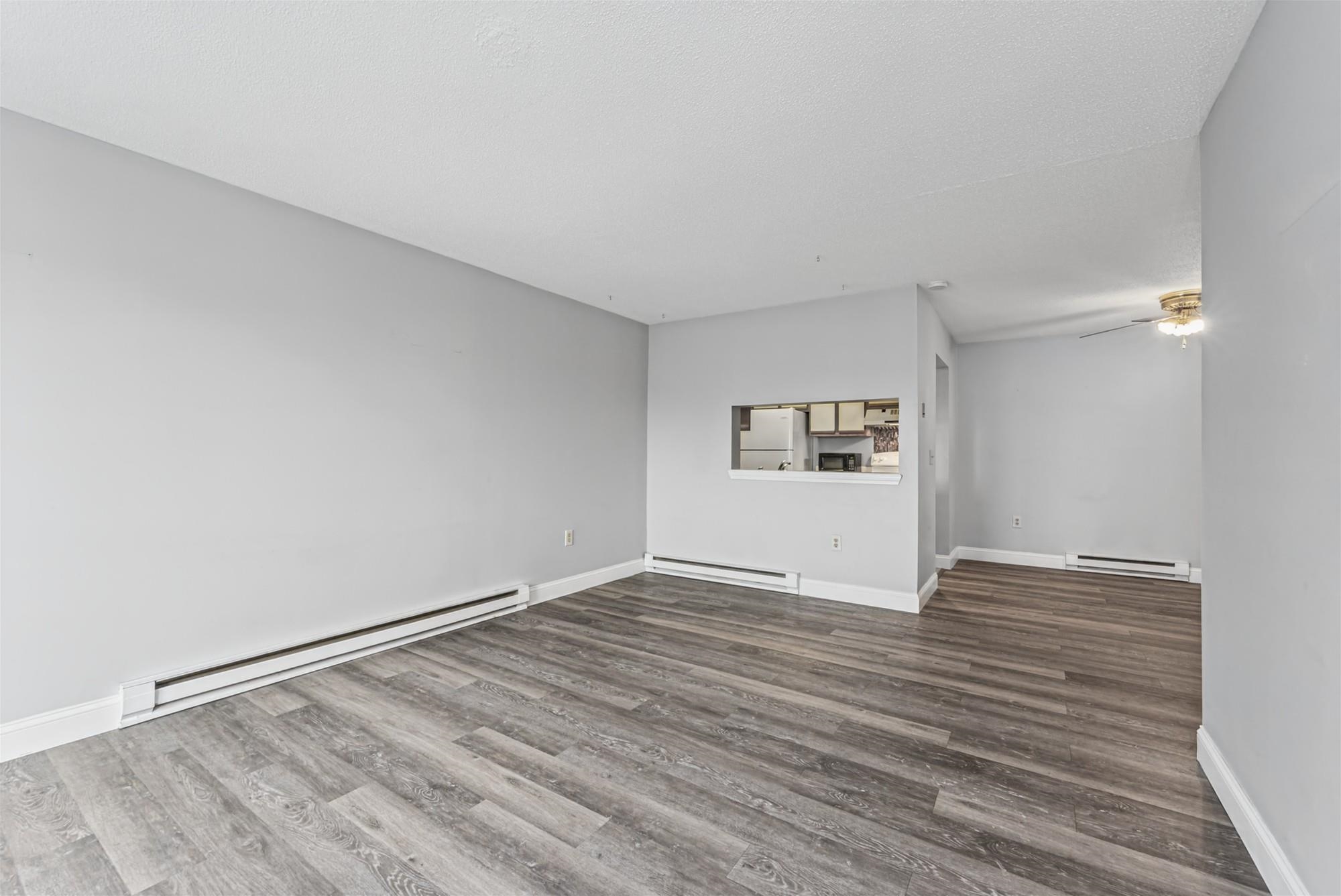 Vacant living room with gray plank flooring, baseboard heaters, and a kitchen pass-through.