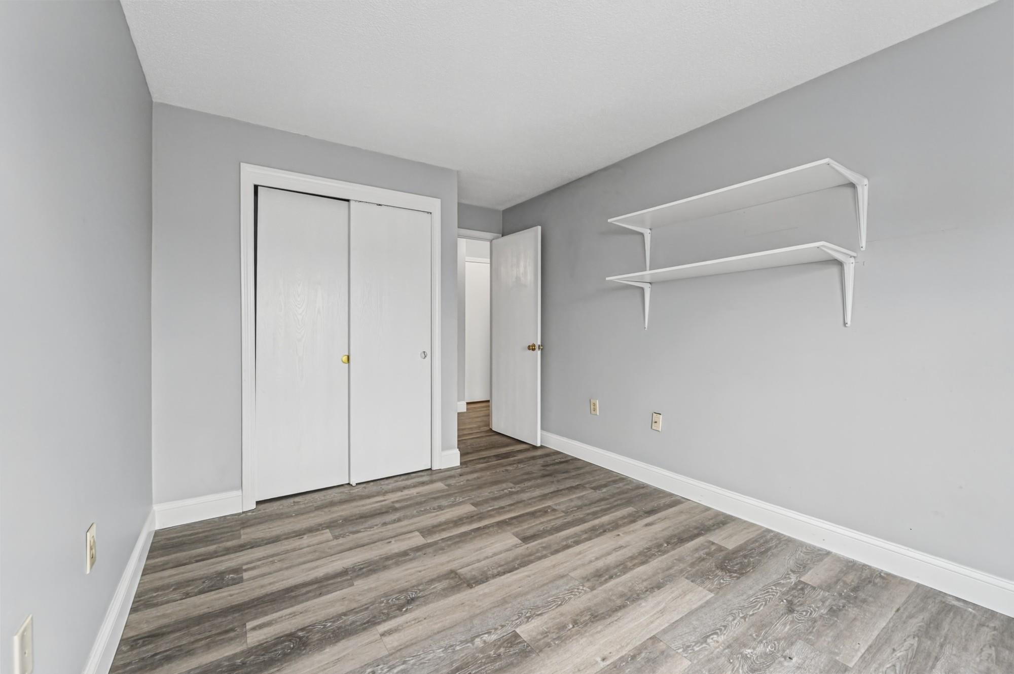 Empty room with gray walls, wood-look flooring, sliding closet doors, and two wall shelves.