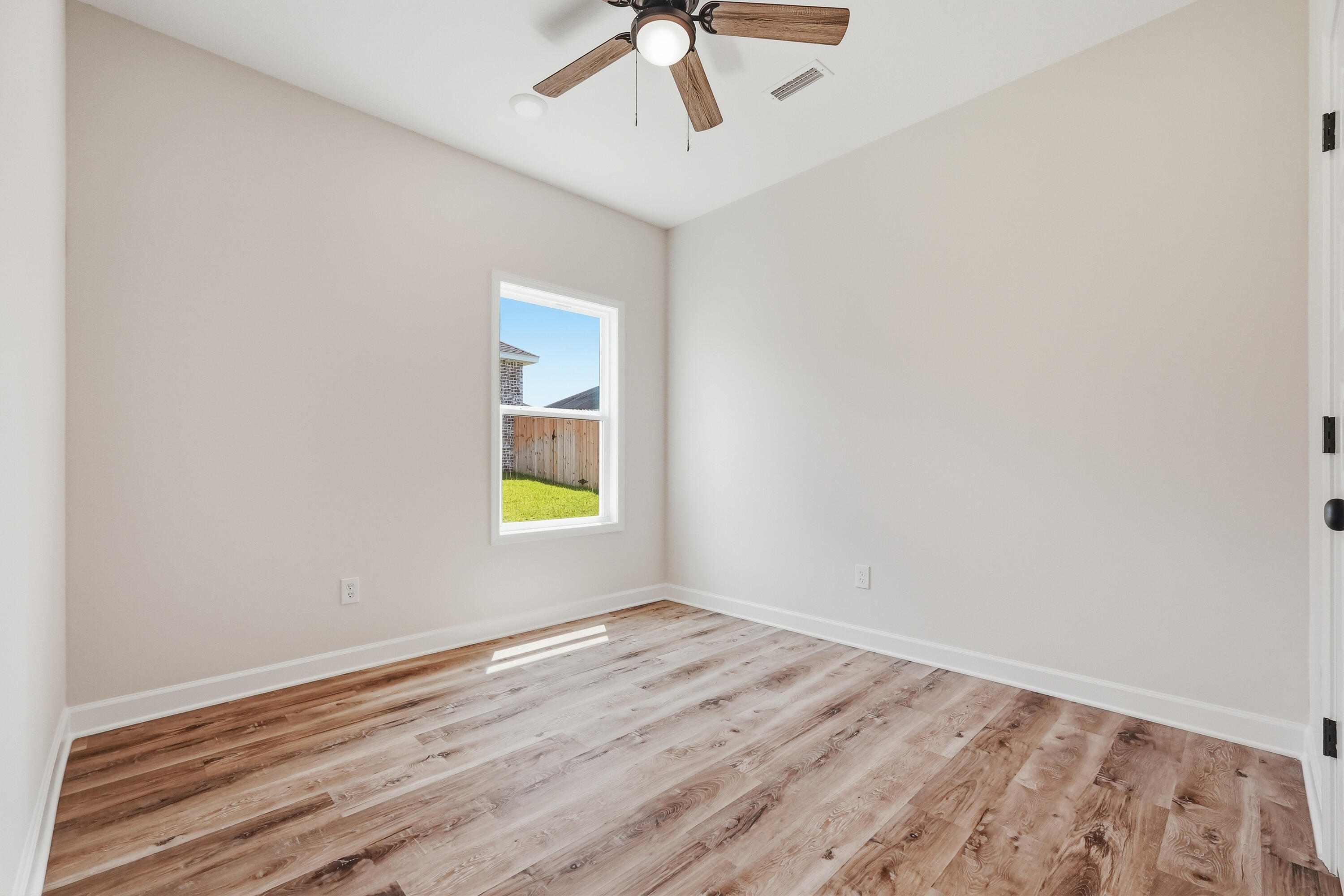 Empty room with wood-look flooring, a window overlooking a fenced yard, and a ceiling fan.