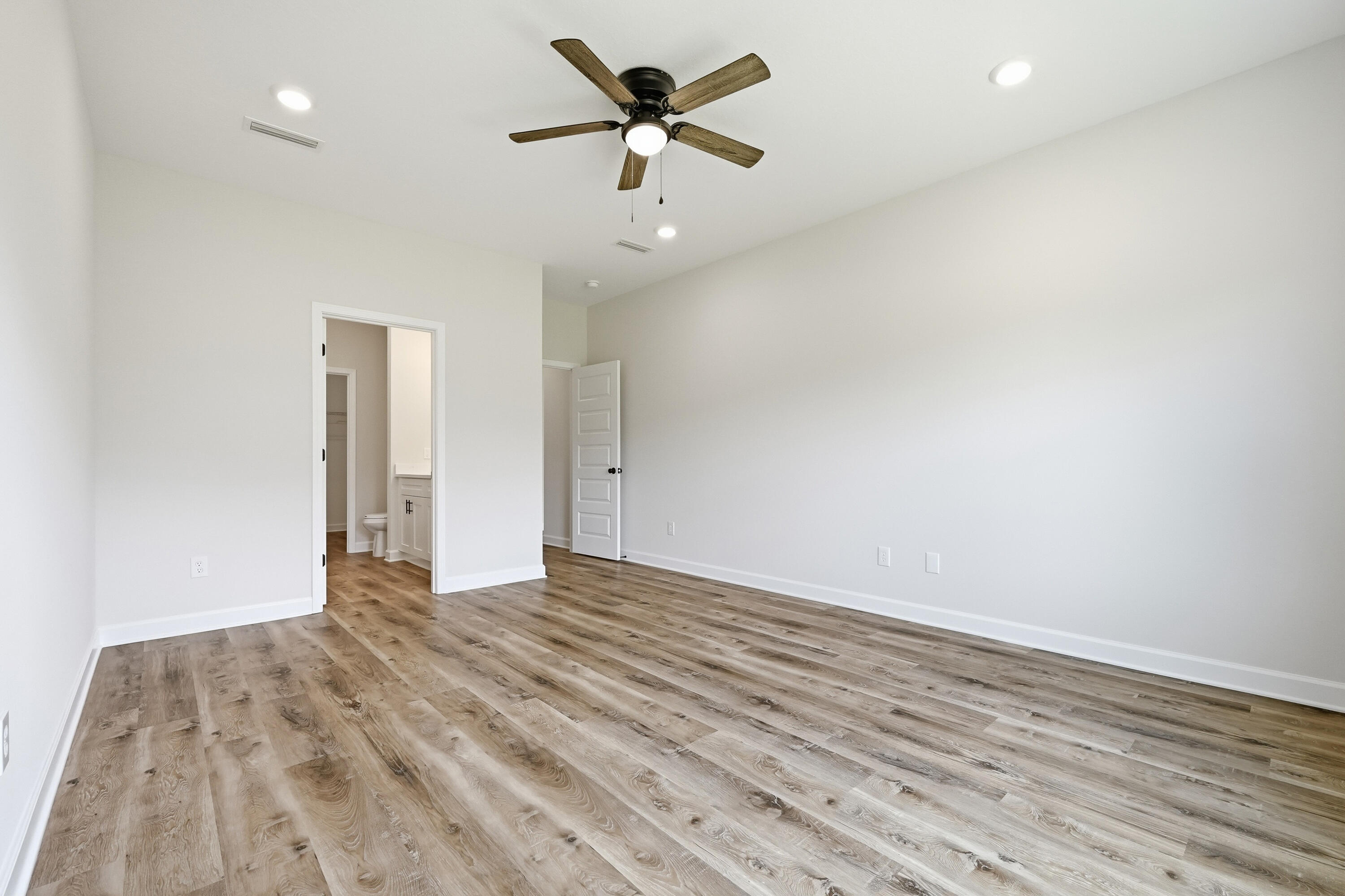 Vacant bedroom with wood-look flooring, a ceiling fan, and an open bathroom doorway.