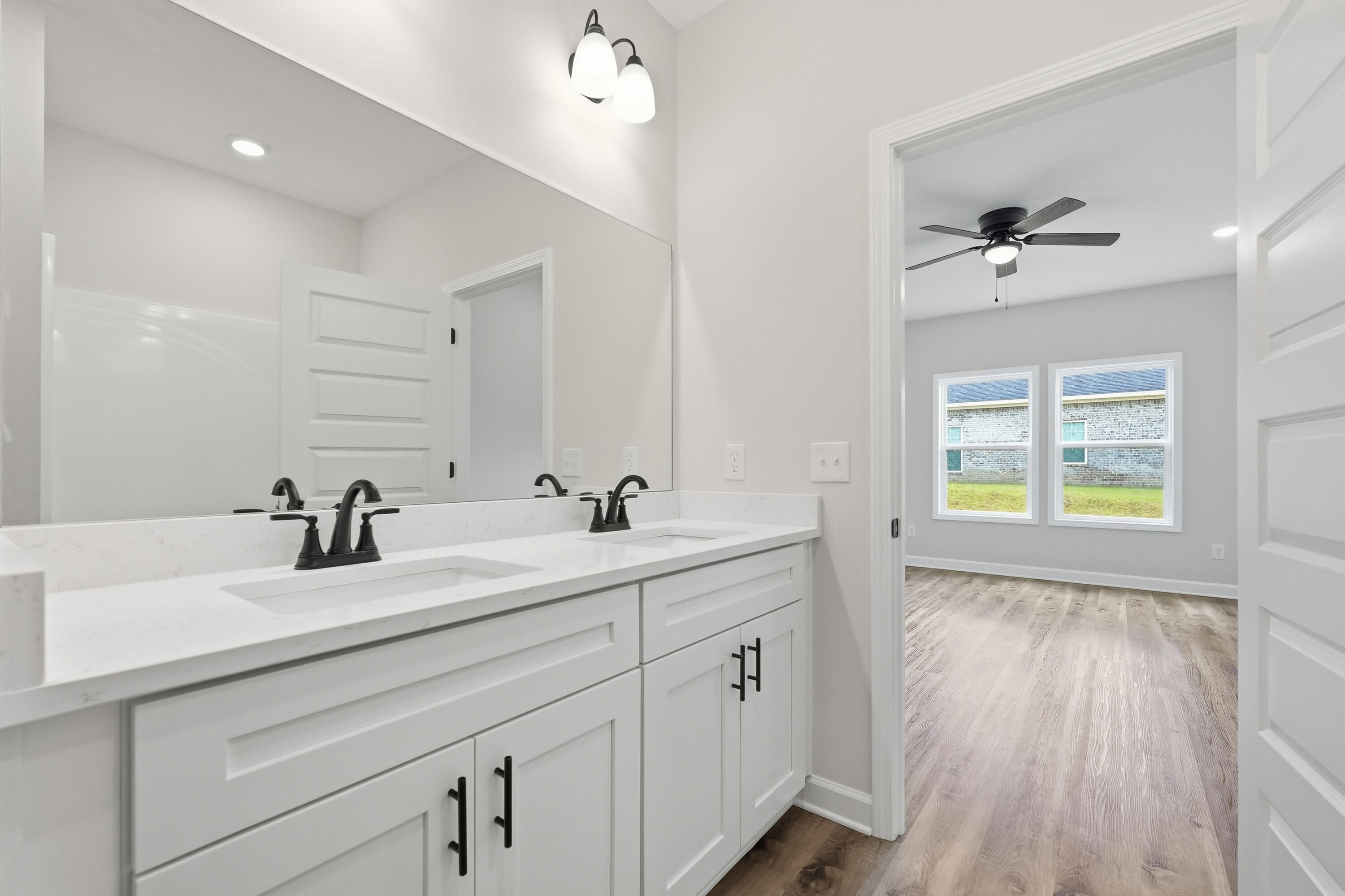 Bathroom with a white double-sink vanity and black faucets, opening to an empty room with two windows.