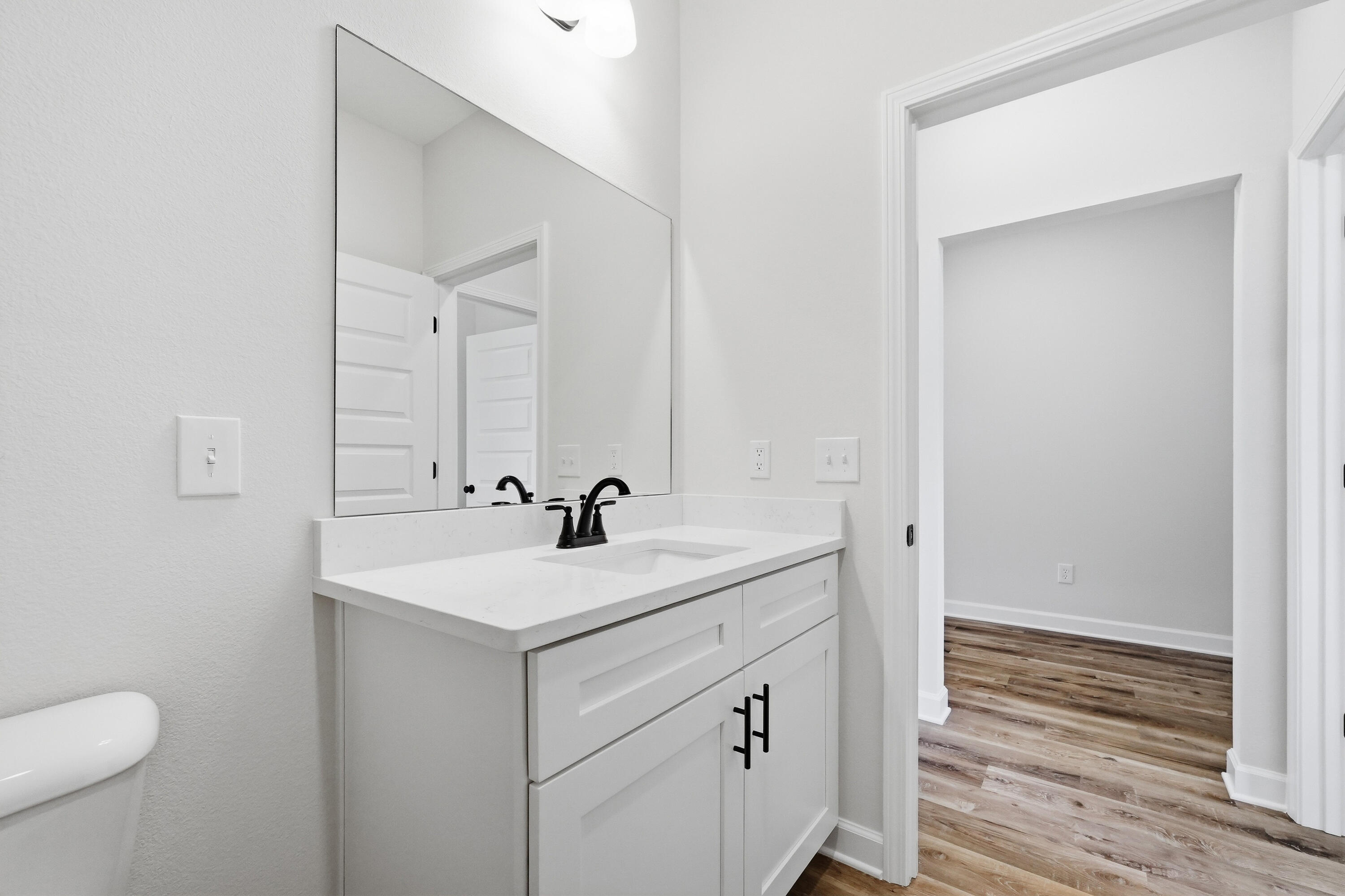 Bathroom vanity with a large mirror, white cabinetry, and black faucet beside an open doorway.