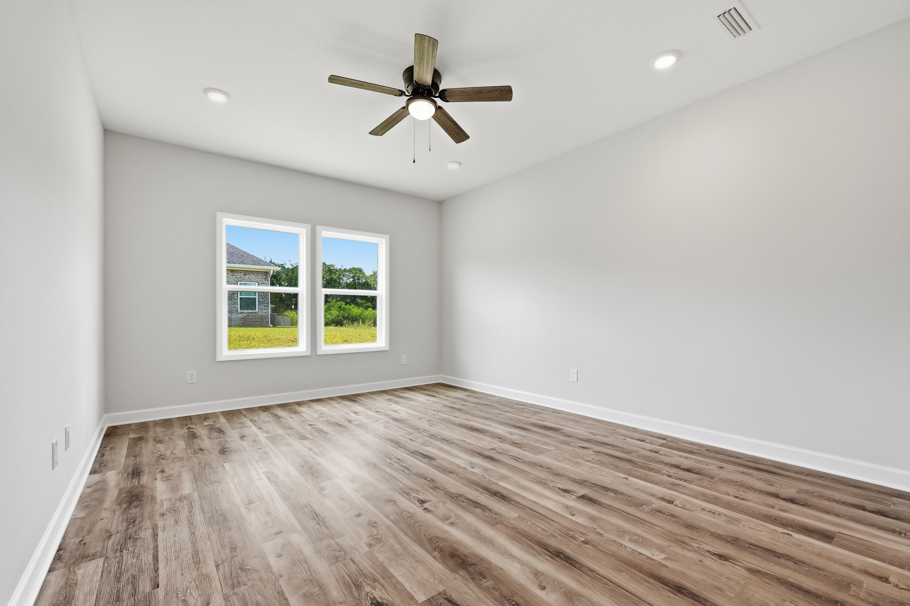 Empty bedroom with wood-look flooring, a ceiling fan, and two windows overlooking the yard.