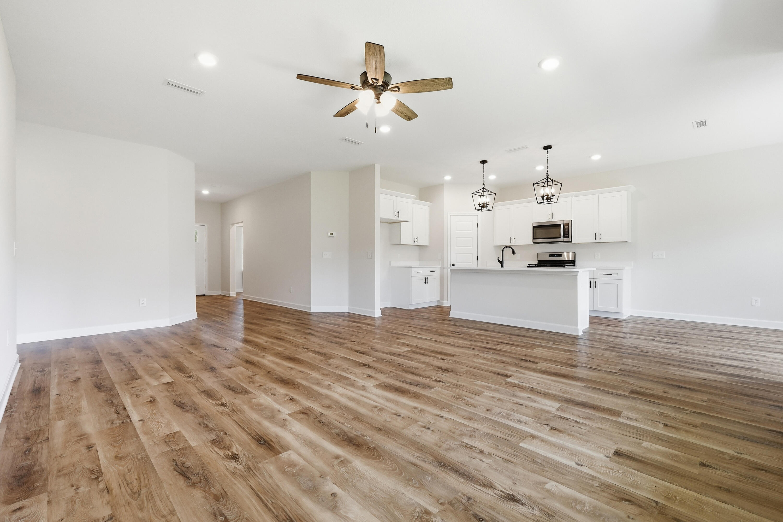 Open living area with wood-look flooring, leading to a white kitchen with an island and ceiling fan.