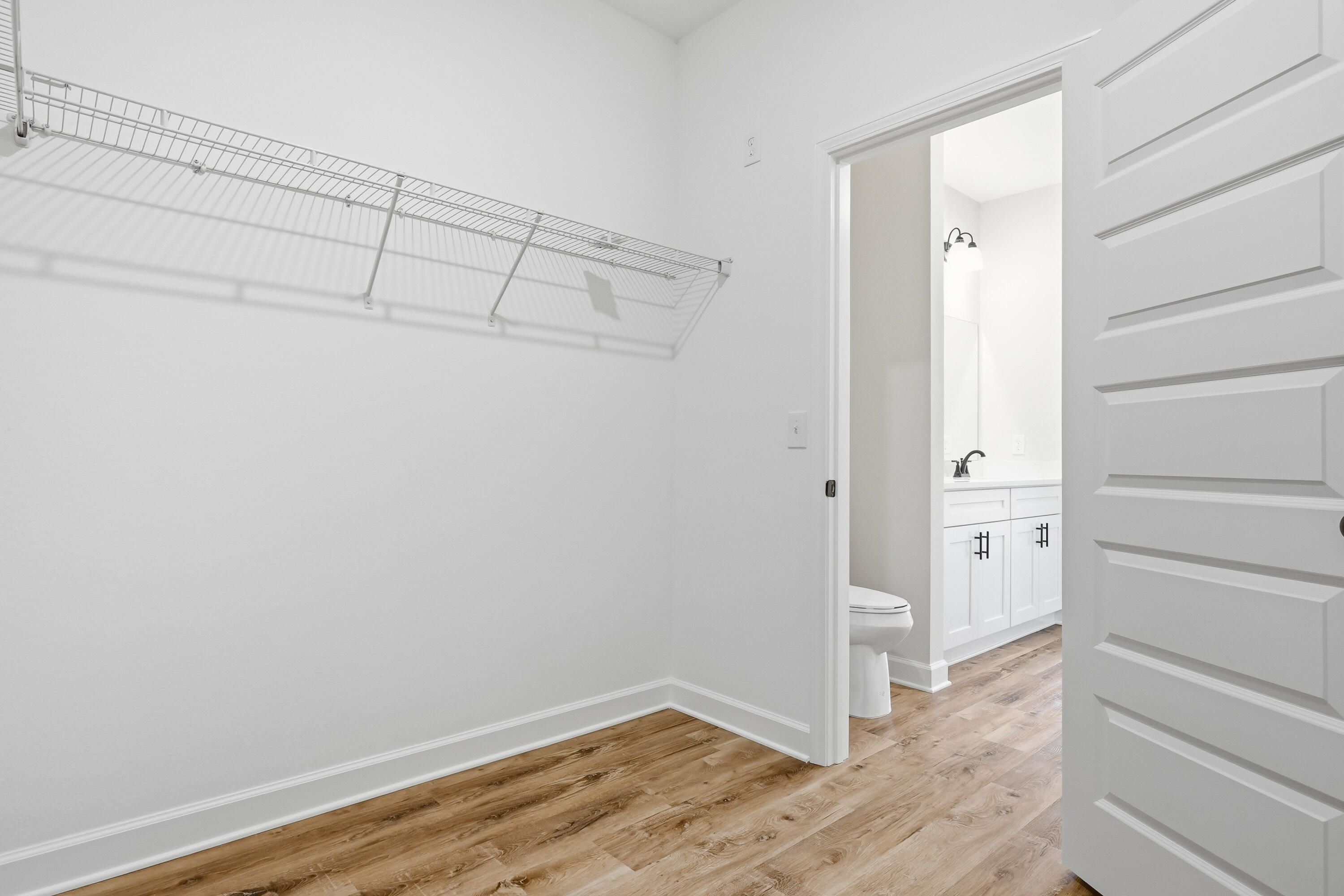 Walk-in closet with wire shelving and wood-look floors opening into a bathroom with a white vanity.