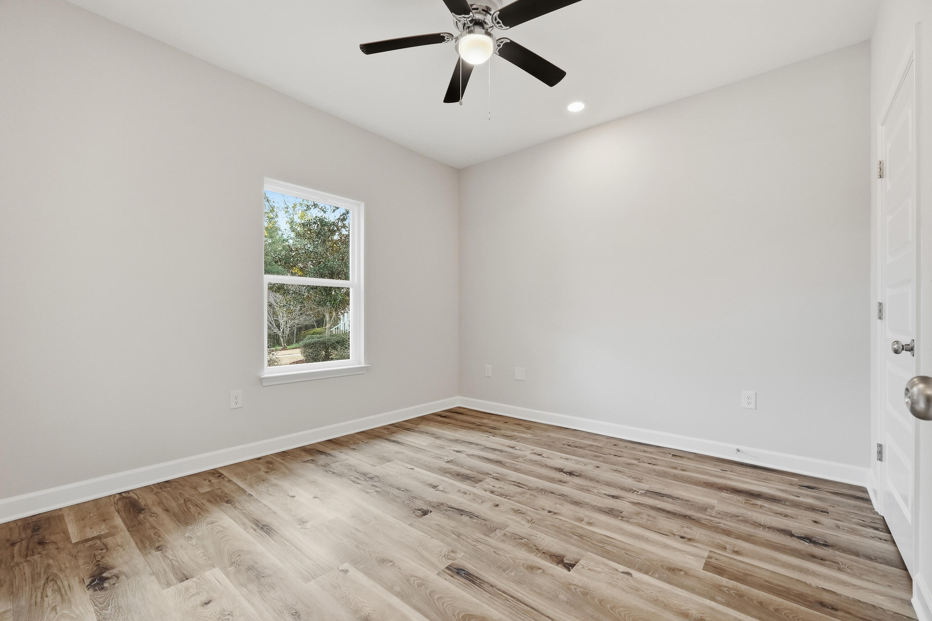 Vacant room with wood-look flooring, a ceiling fan, and a window overlooking greenery.