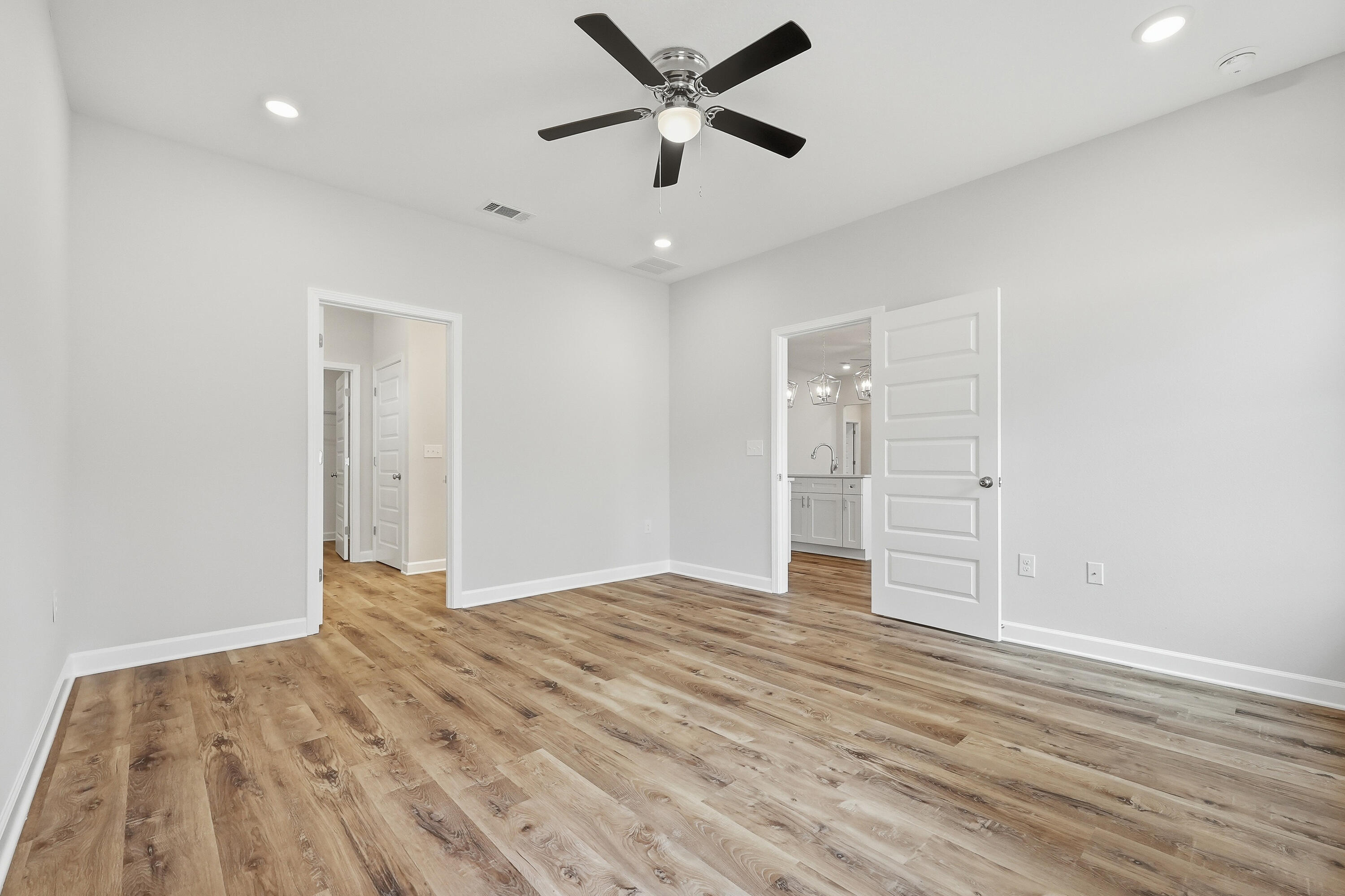 Empty bedroom with wide-plank wood-look floors and a ceiling fan, opening to a bathroom vanity.