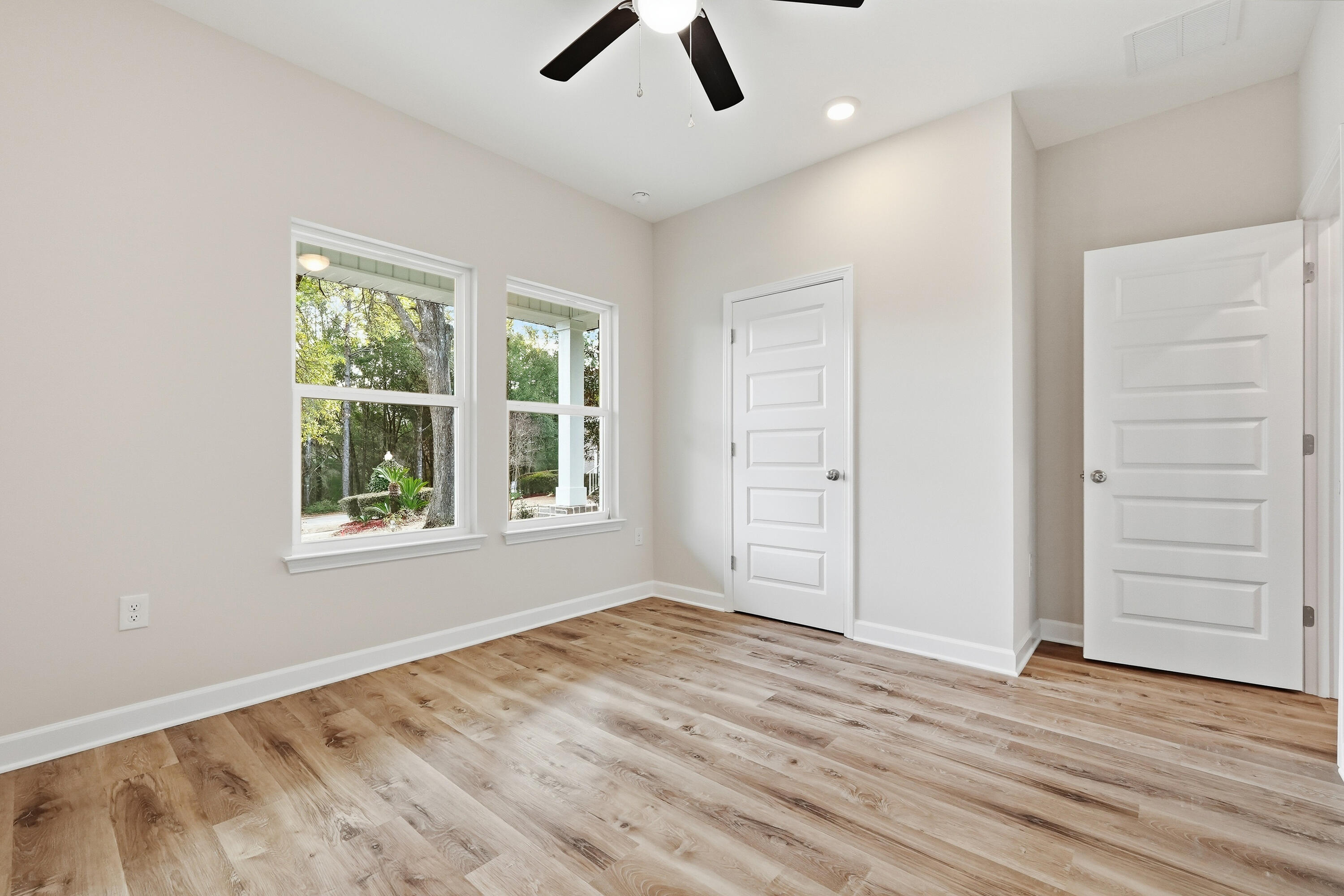 Empty bedroom with wood flooring, ceiling fan, and two windows overlooking trees.
