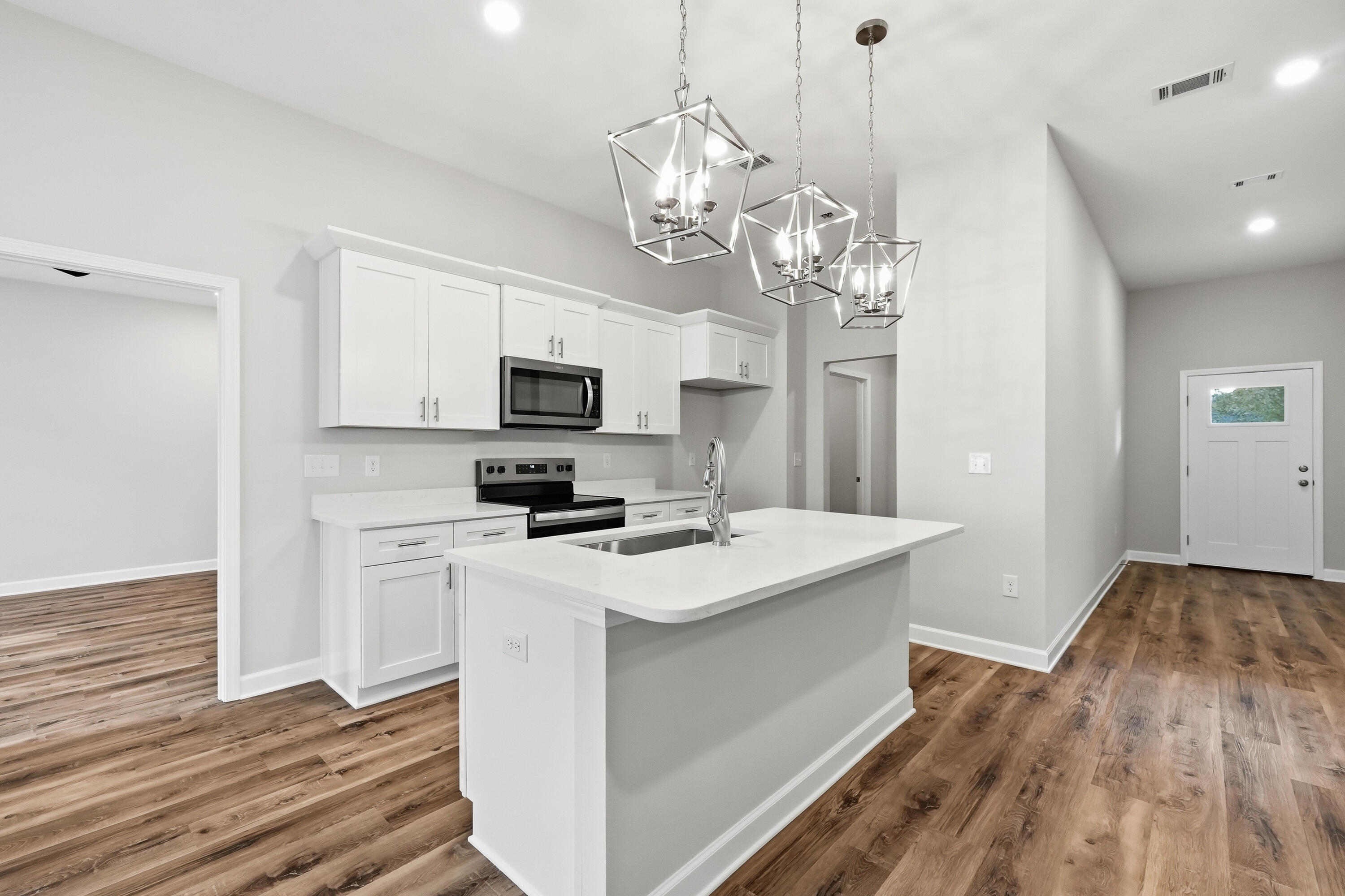 Vacant kitchen with a large white island, stainless steel appliances, geometric pendants, and wood-look floors.