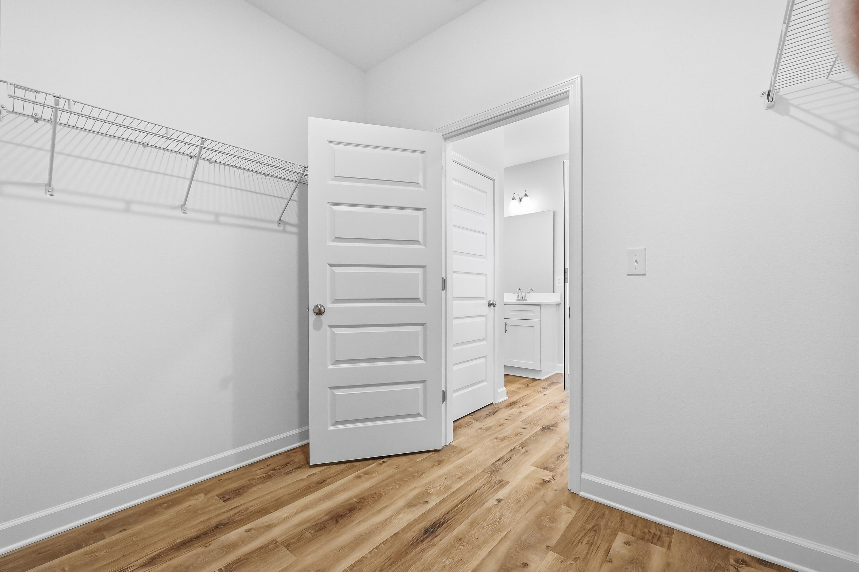 Empty walk-in closet with wire shelving and wood-look flooring, opening to a bathroom vanity.