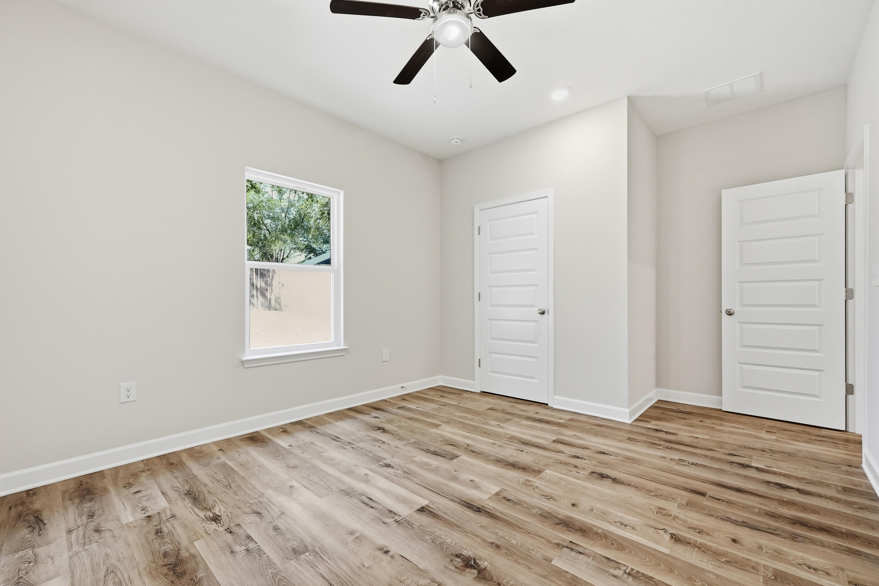 Empty room with wood-look flooring, a ceiling fan, window, and two white doors.