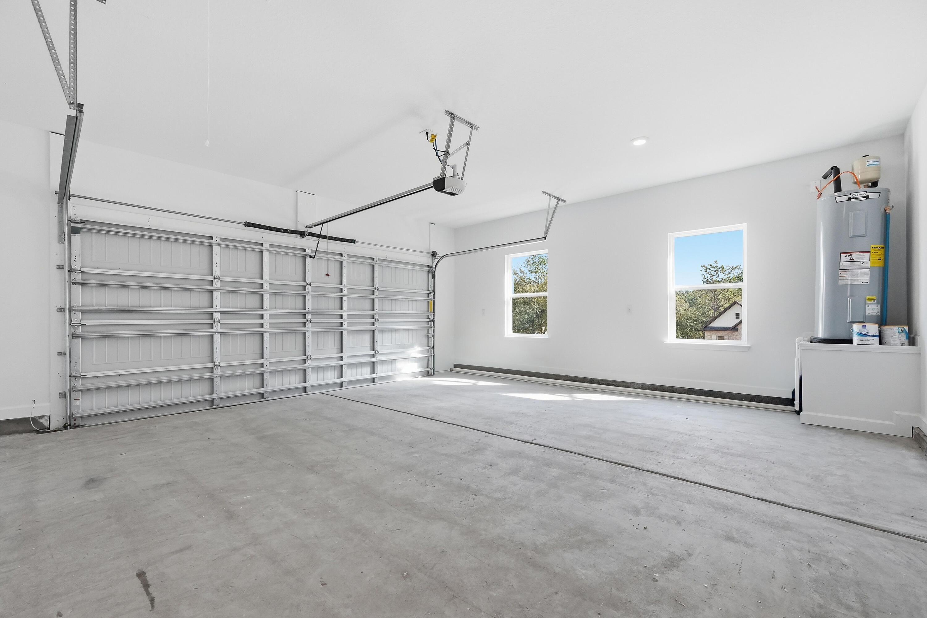 Empty garage with a wide overhead door, concrete floor, two windows, and a water heater.