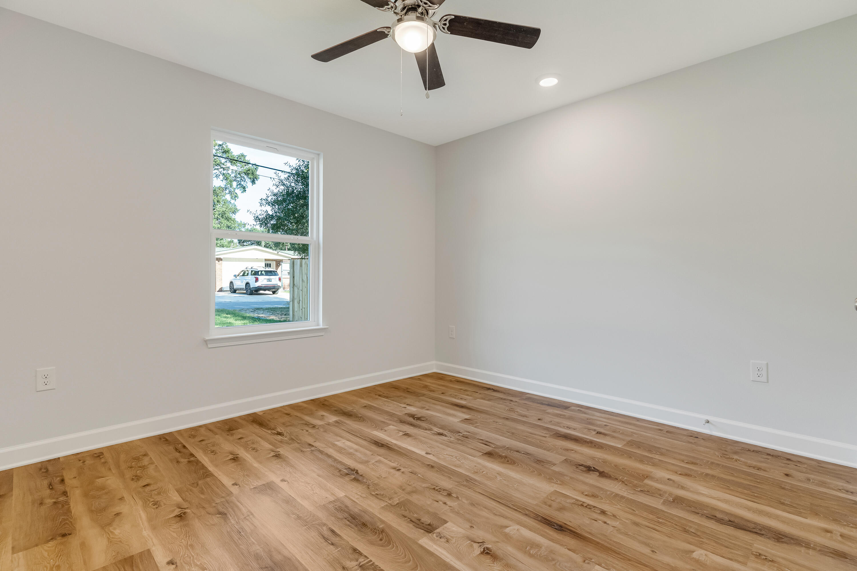 Empty room with wide-plank wood flooring, a ceiling fan, and a window overlooking a neighboring home.