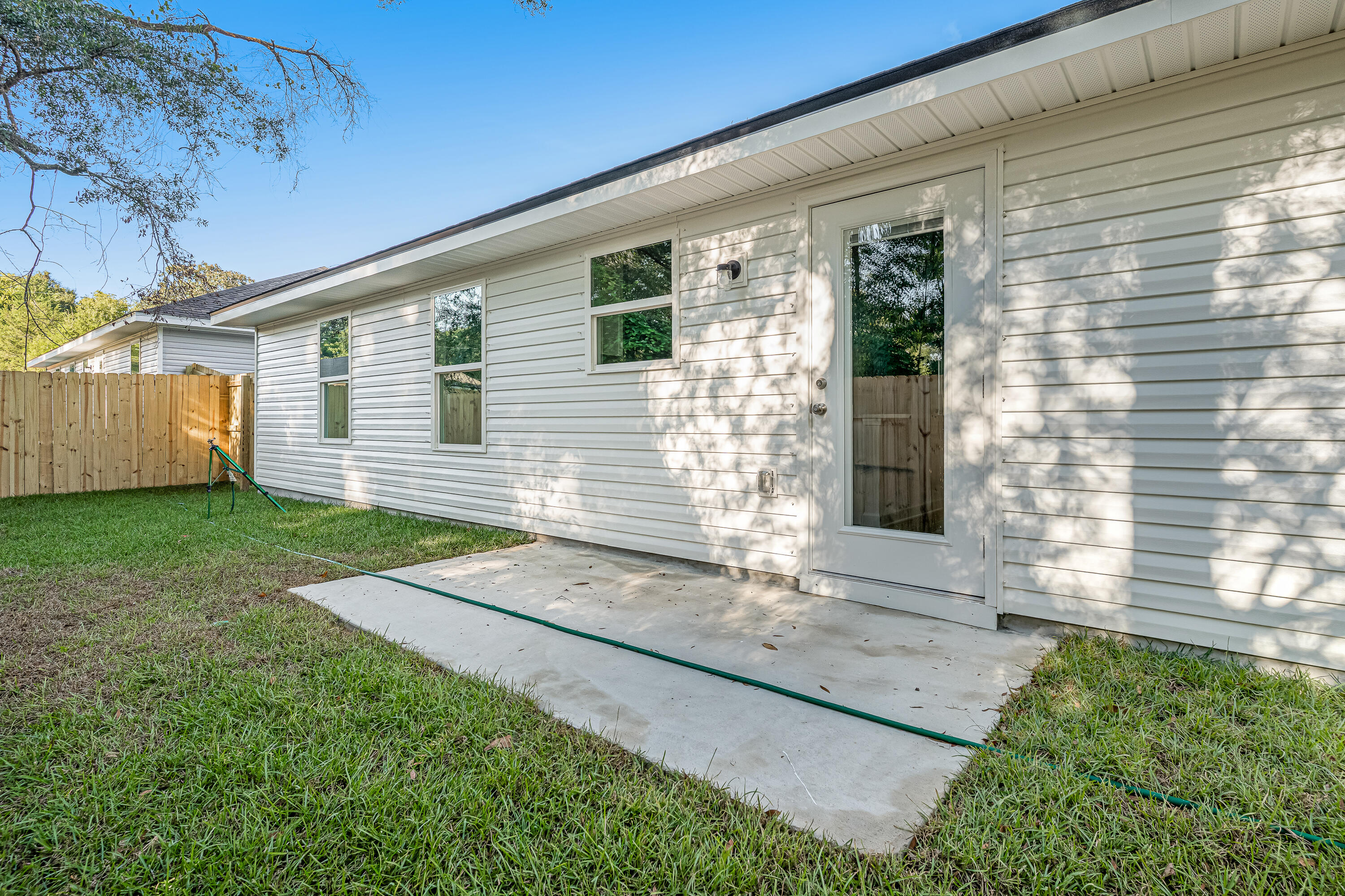 Rear of a white-sided house with a glass-panel door, concrete patio, and fenced grassy yard.