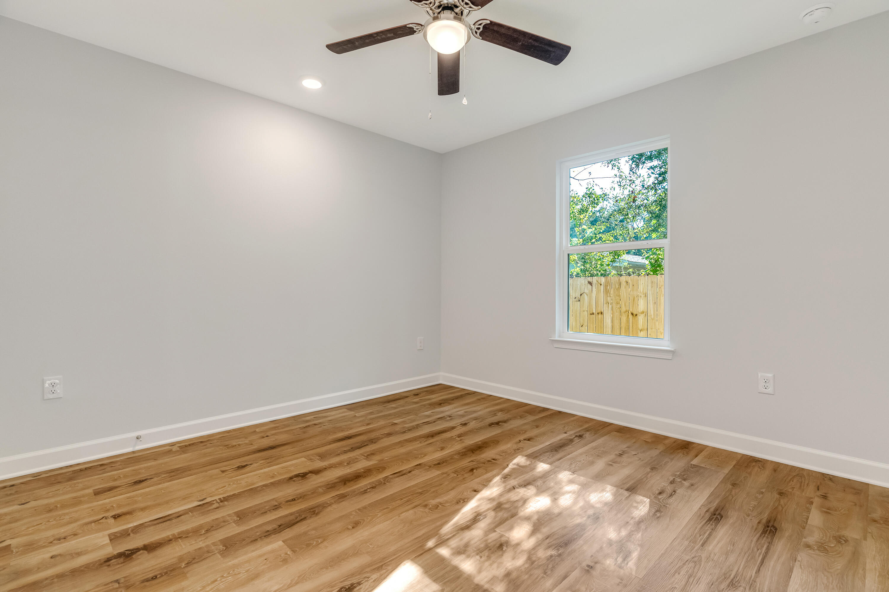 Empty room with warm wood flooring, ceiling fan, and a window overlooking trees and a wooden fence.
