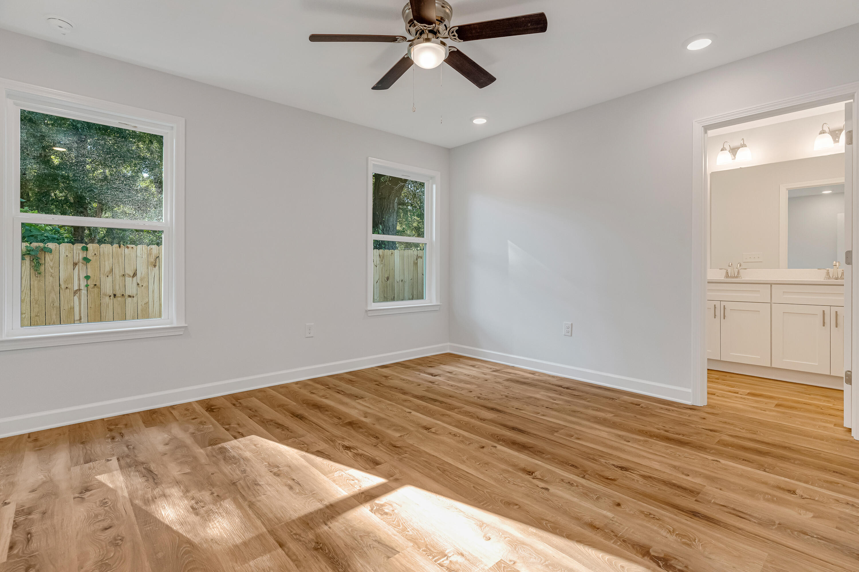 Vacant bedroom with wood flooring, ceiling fan, and doorway to a bathroom with double vanity.
