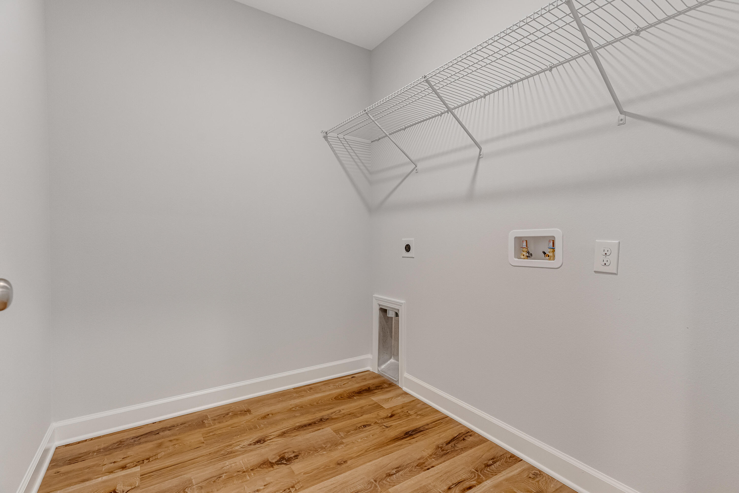 Empty laundry room with a wire shelf, washer hookups, and warm wood-look flooring.