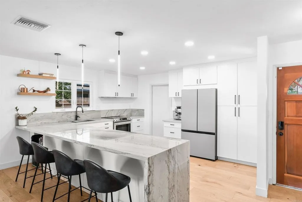 Bright white kitchen with a marble waterfall island, black stools, and stainless steel refrigerator.