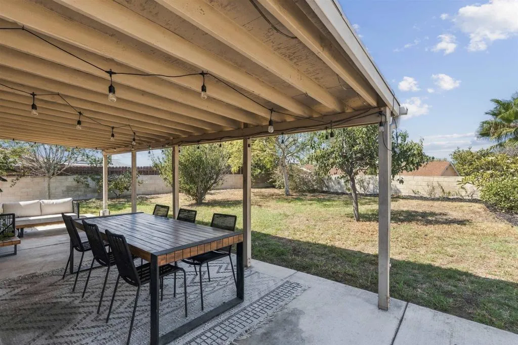 Covered backyard patio with outdoor dining table, string lights, and a dry lawn beyond.