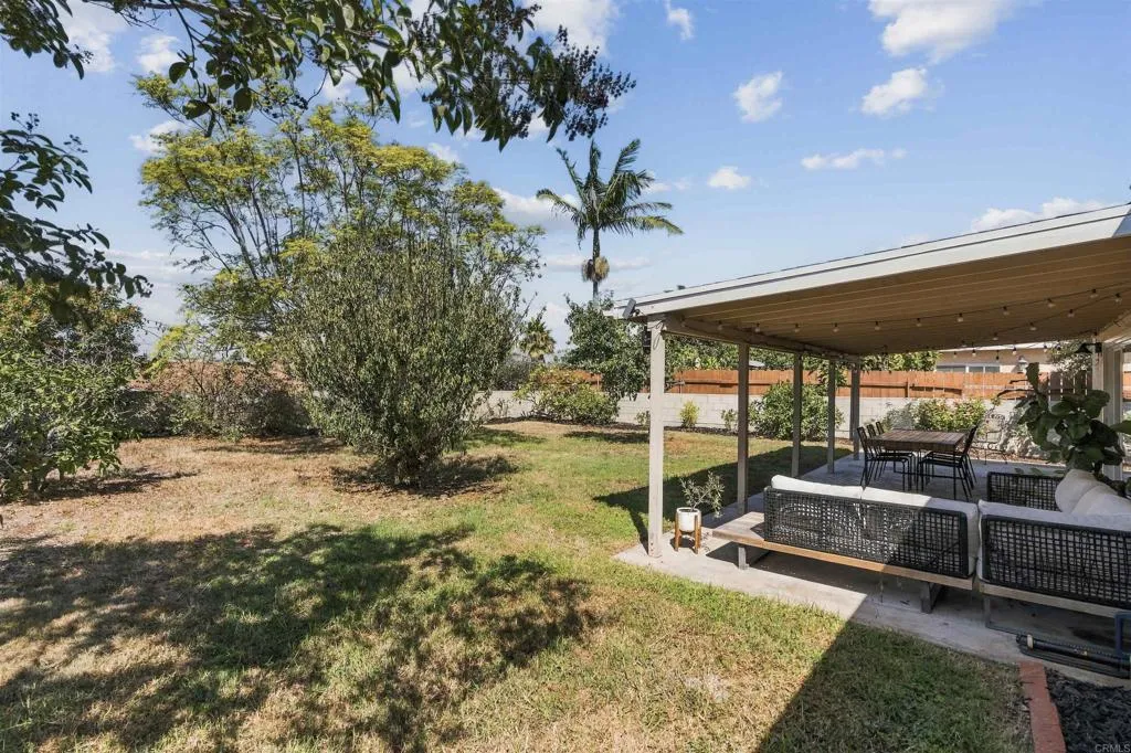 Backyard lawn with mature trees beside a covered patio with outdoor seating and a dining table.