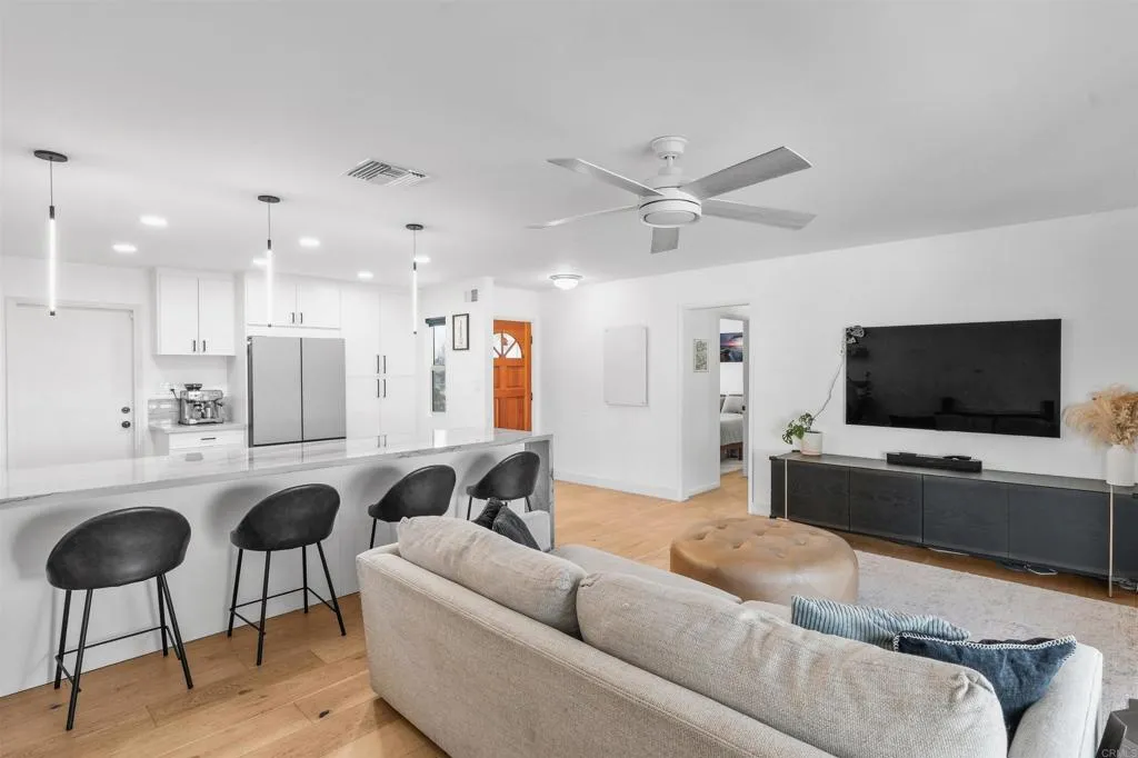 Open-plan living room and kitchen with a light sofa, wall-mounted television, and island with bar stools.