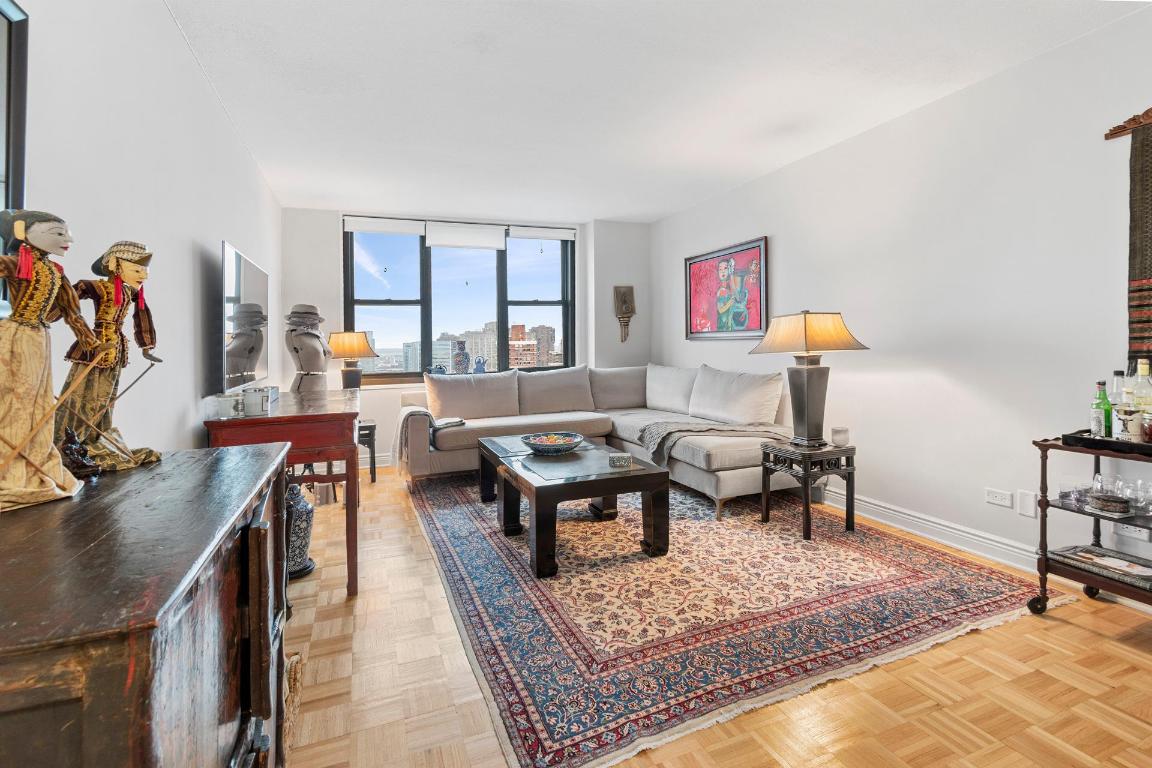 Living room with a light sectional sofa, patterned rug, and large windows overlooking city buildings.