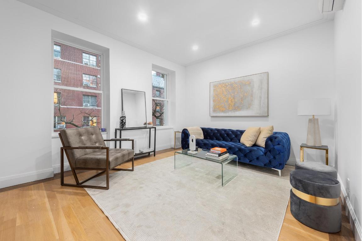 Living room with a blue tufted sofa, gray armchair, and two large windows overlooking a brick building.