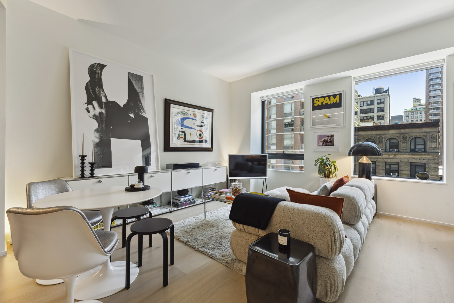 Modern living room with a rounded white sofa, dining table, and large windows overlooking city buildings.