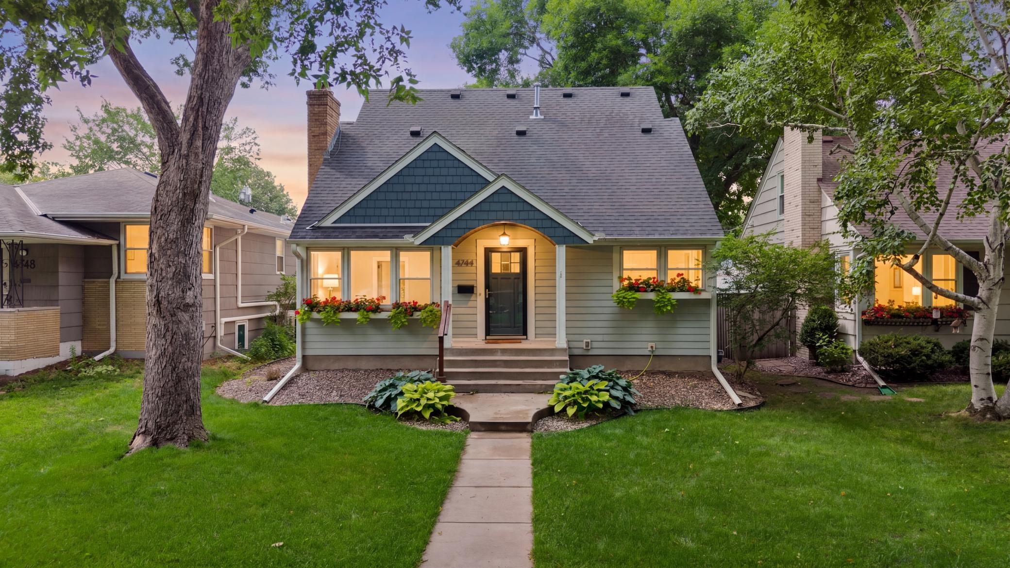 Gray Minneapolis home with blue gables, flower boxes, and a tree-lined front yard at dusk.