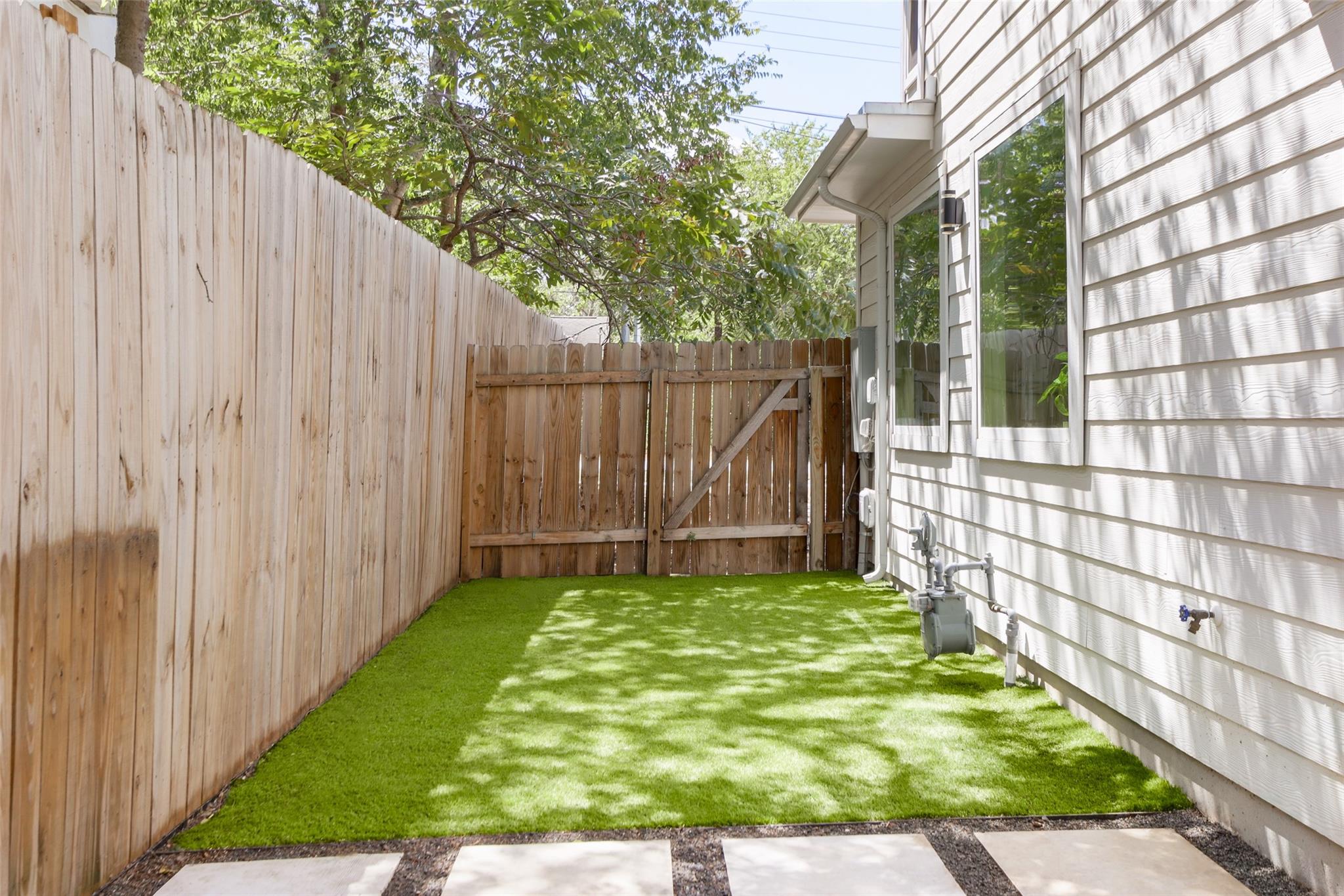 Fenced side yard with green lawn, wooden gate, and patio pavers beside a white-sided house.