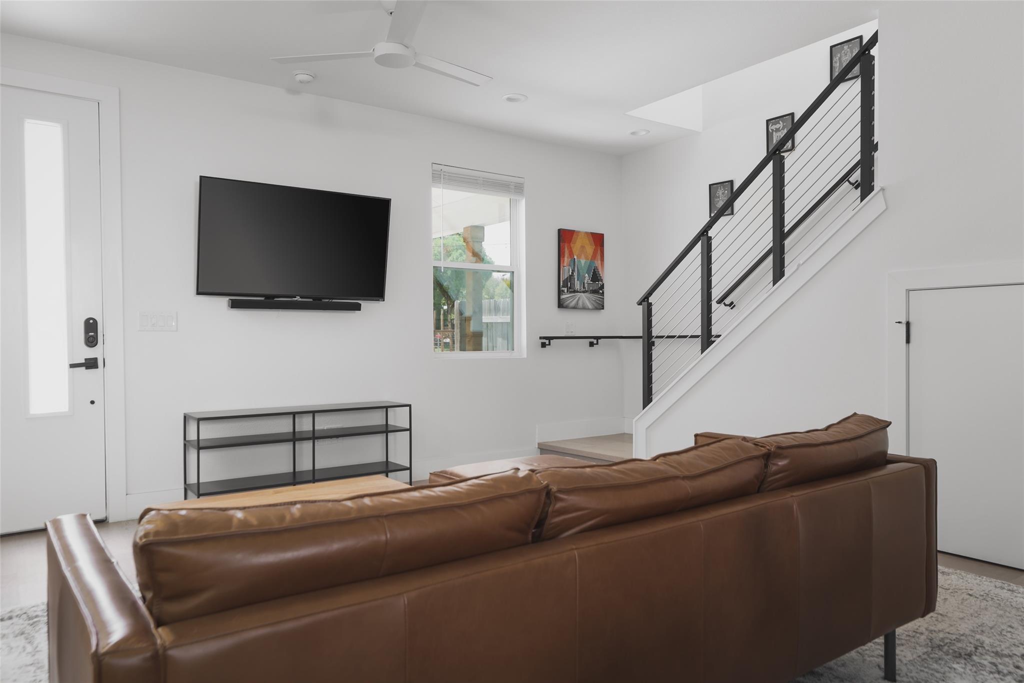 Living room with a brown leather sectional, wall-mounted TV, and staircase with black cable railing.