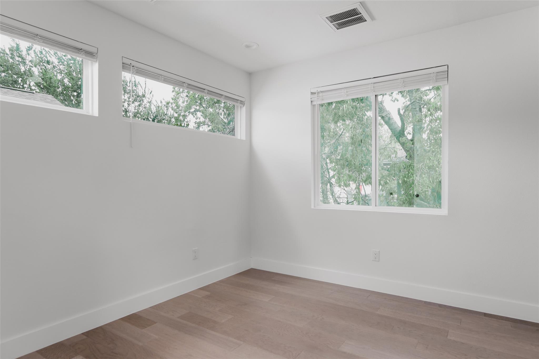 Empty room with light wood flooring and three windows overlooking leafy trees.