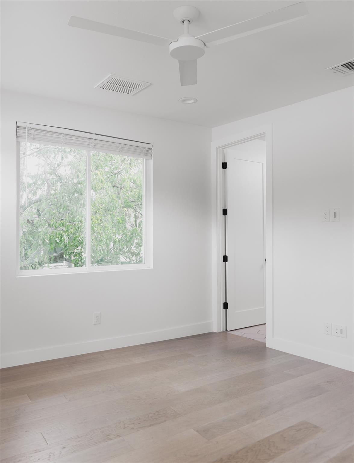 Vacant room with pale wood floors, a ceiling fan, and a large window overlooking leafy greenery.