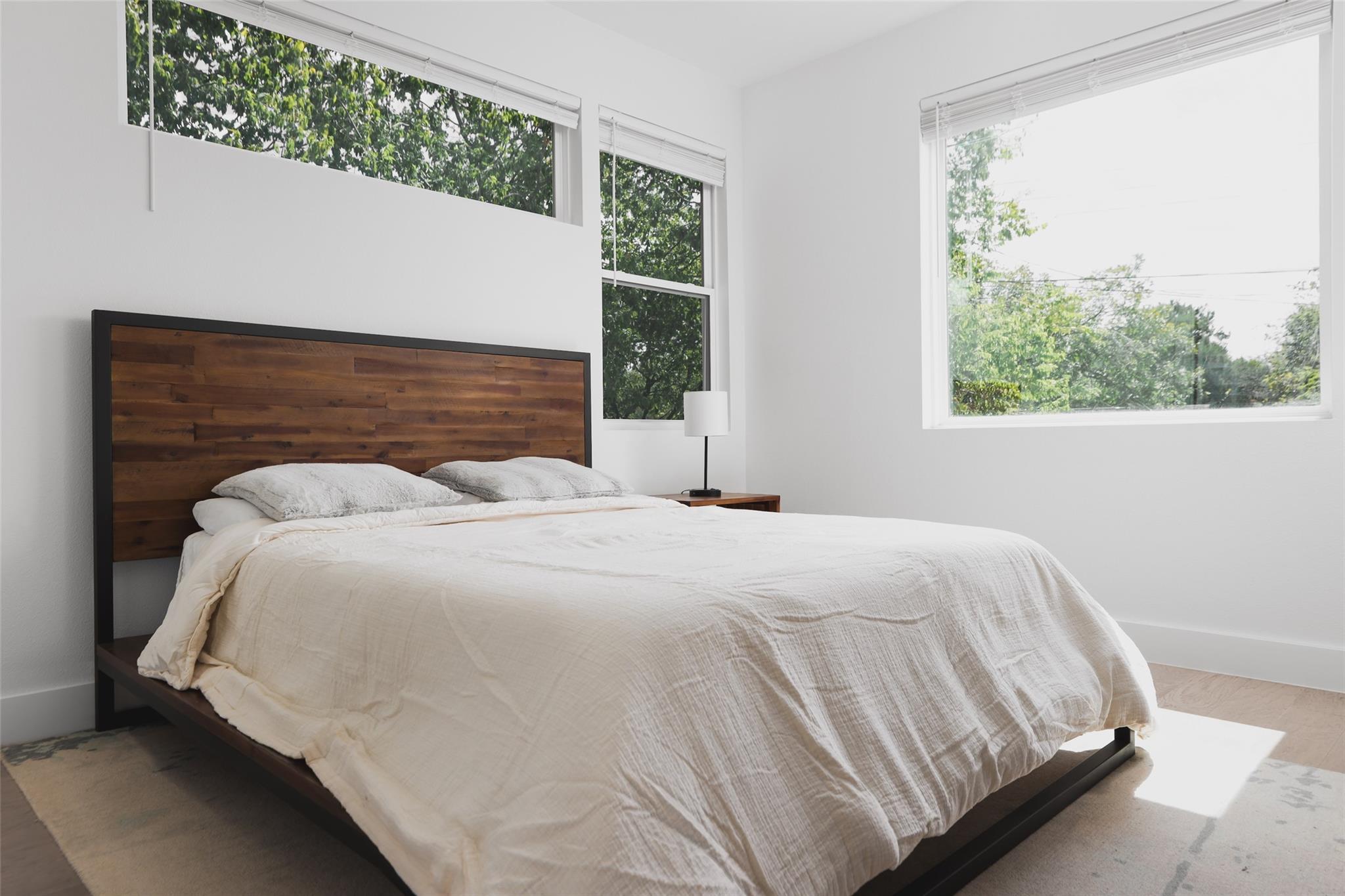 Bright bedroom with a large white bed, wood headboard, and tall windows overlooking trees.