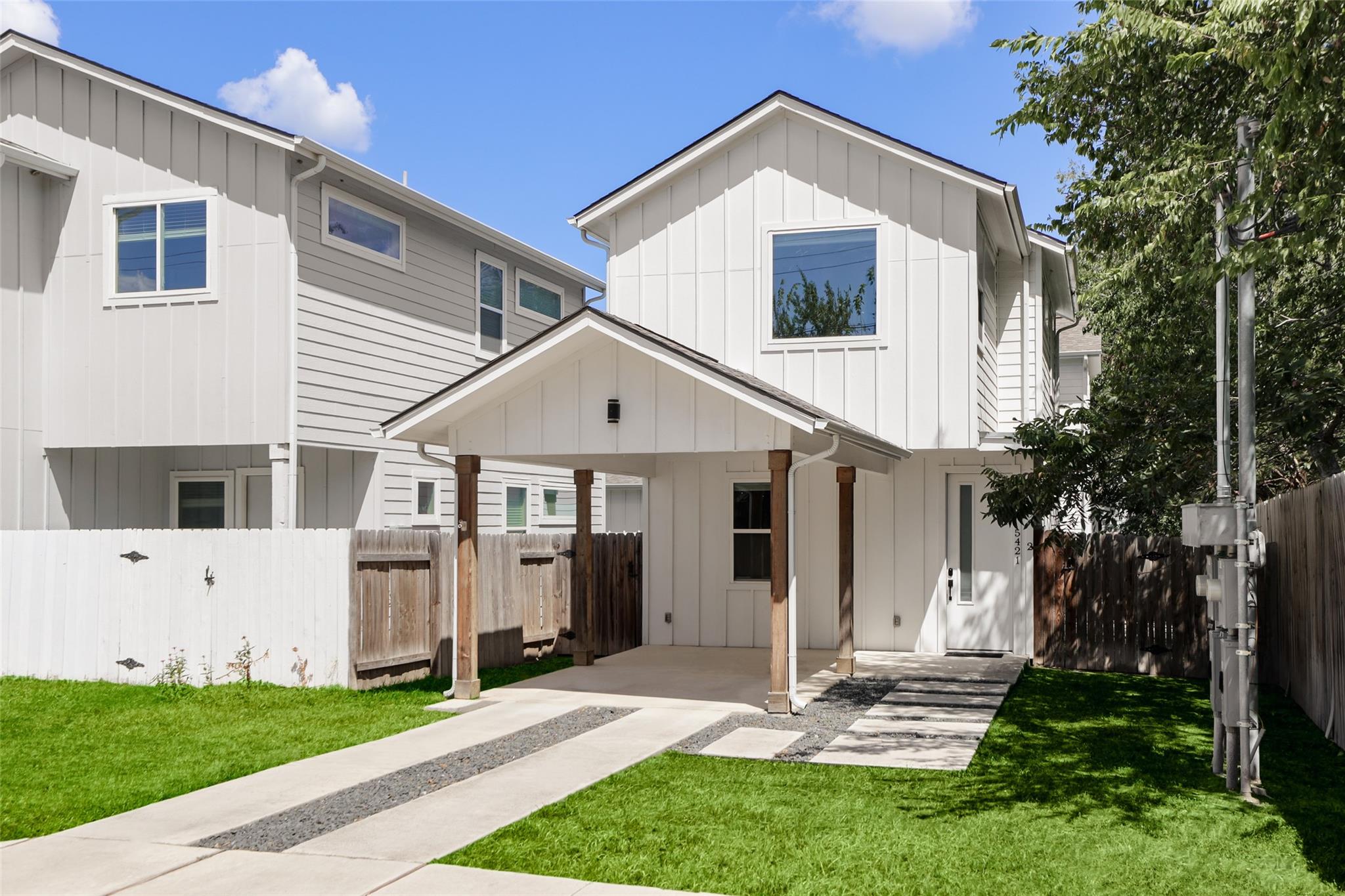 White two-story home with a covered entry, concrete walkways, fenced lawn, and mature trees.