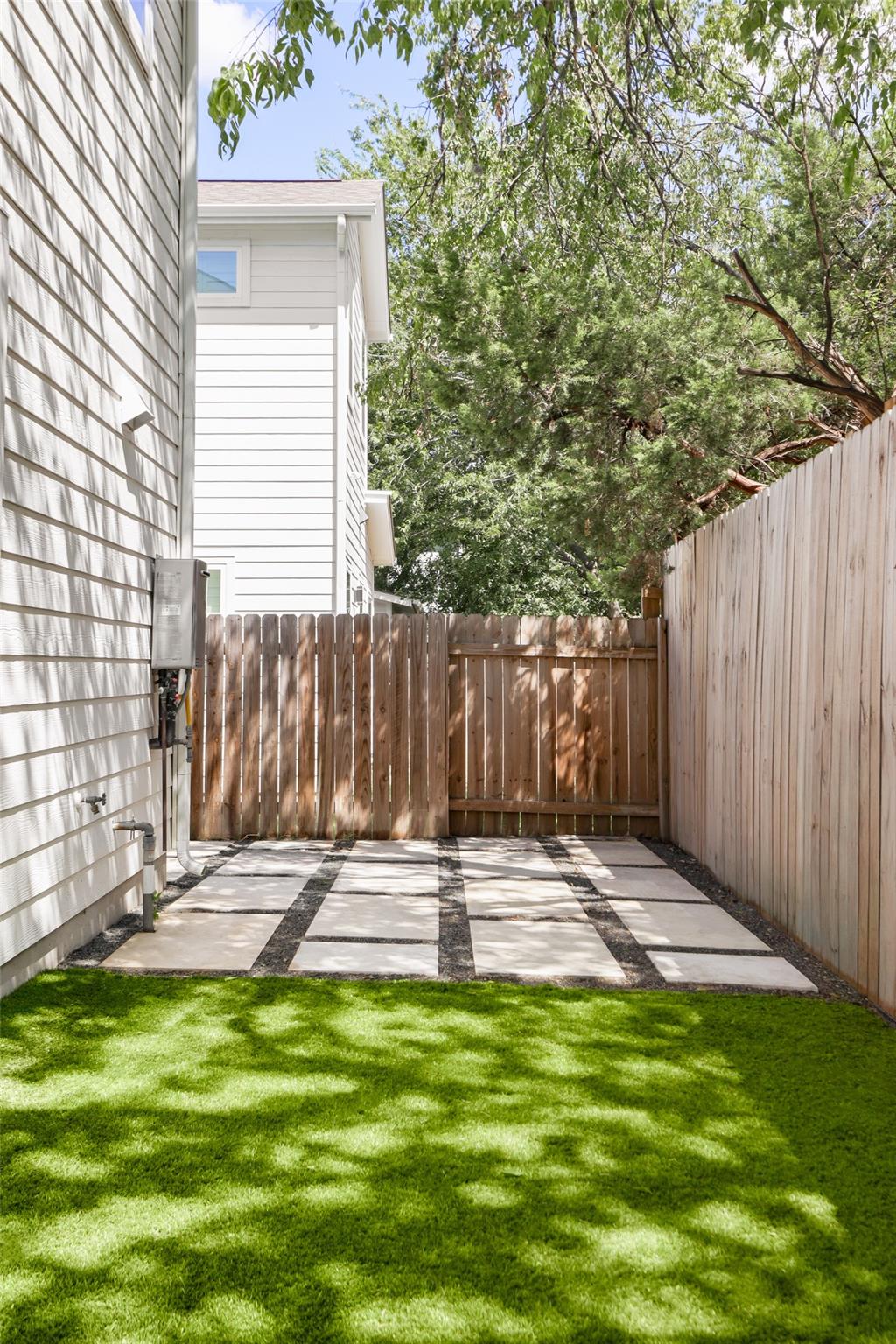 Fenced side yard with artificial turf and a paved patio beneath mature trees.