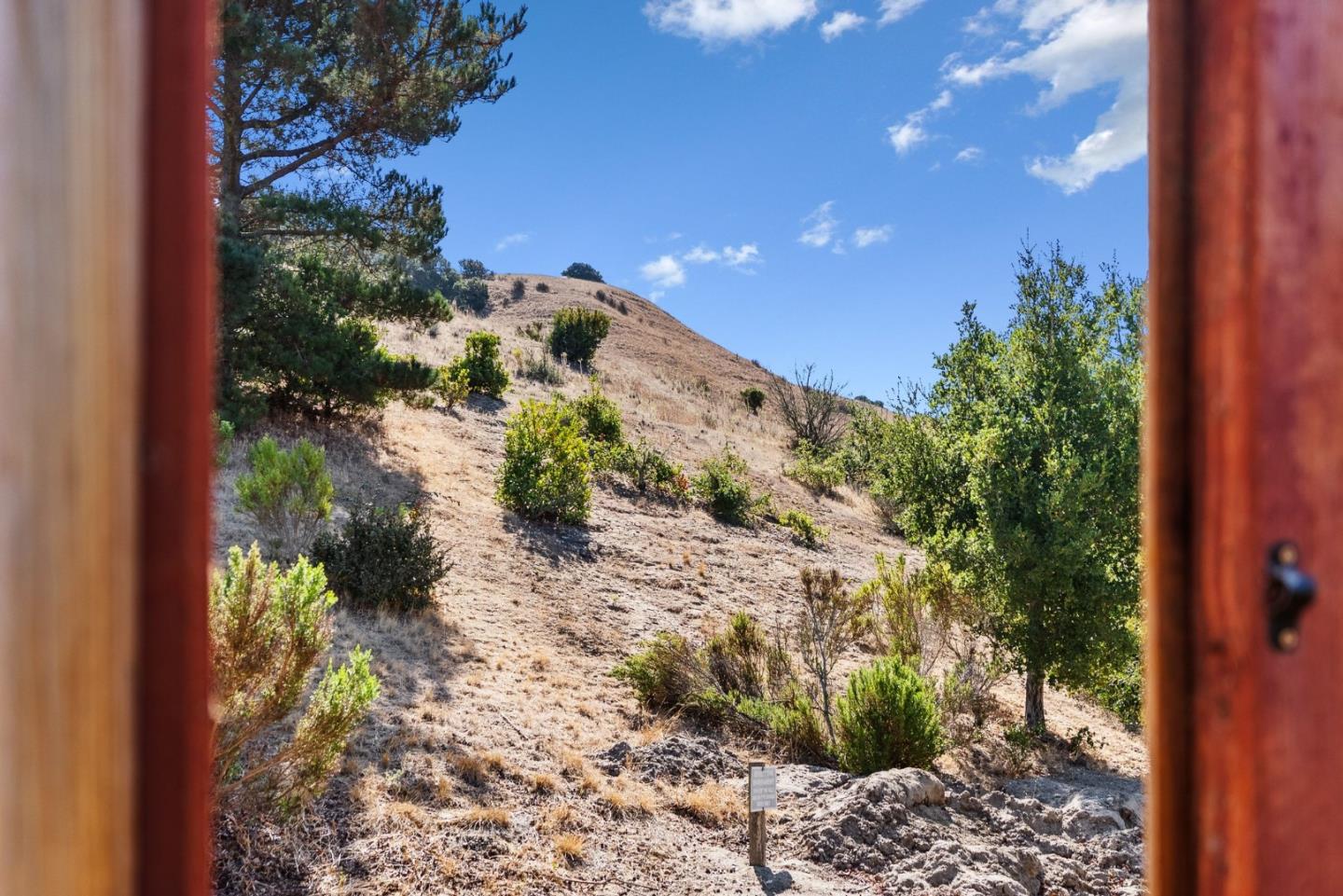Dry, shrub-covered hillside viewed through a wooden doorway, with scattered trees under a blue sky.